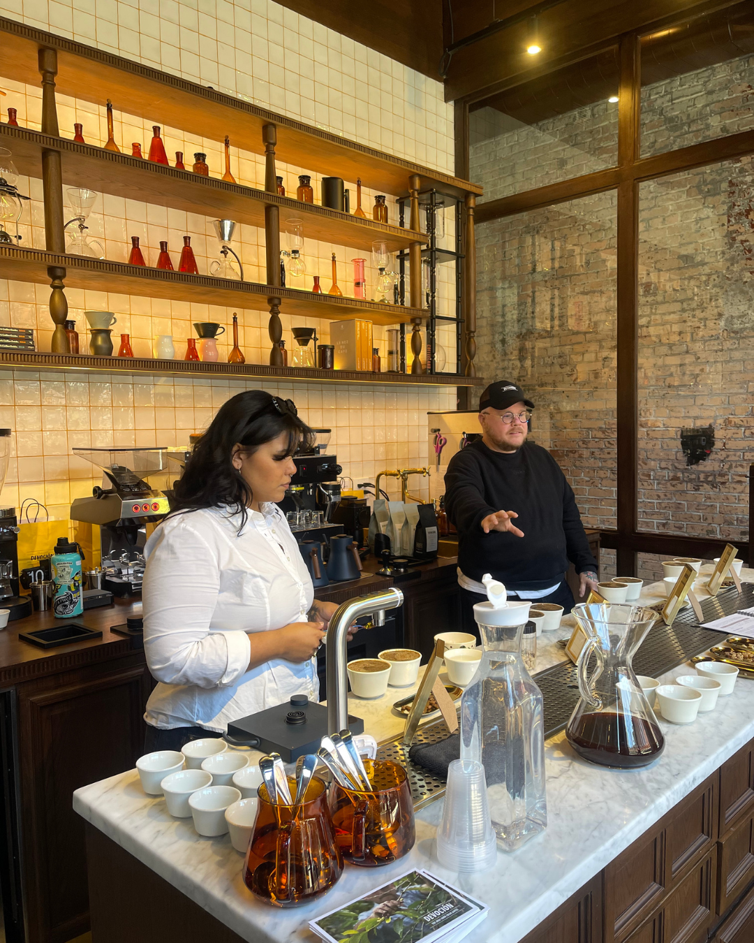 Two people behind a marble café counter with cups, carafes, and coffee brewing equipment