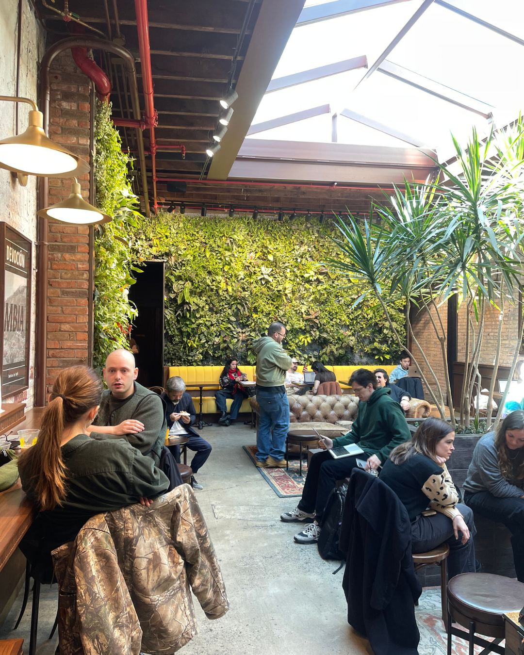 Indoor café lounge with skylight, exposed brick, vertical garden wall, and people socializing at tables