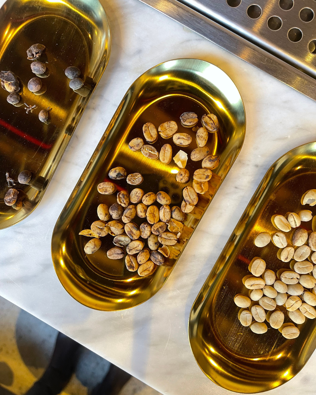 Three golden trays on marble surface showing different stages of coffee beans from raw to roasted