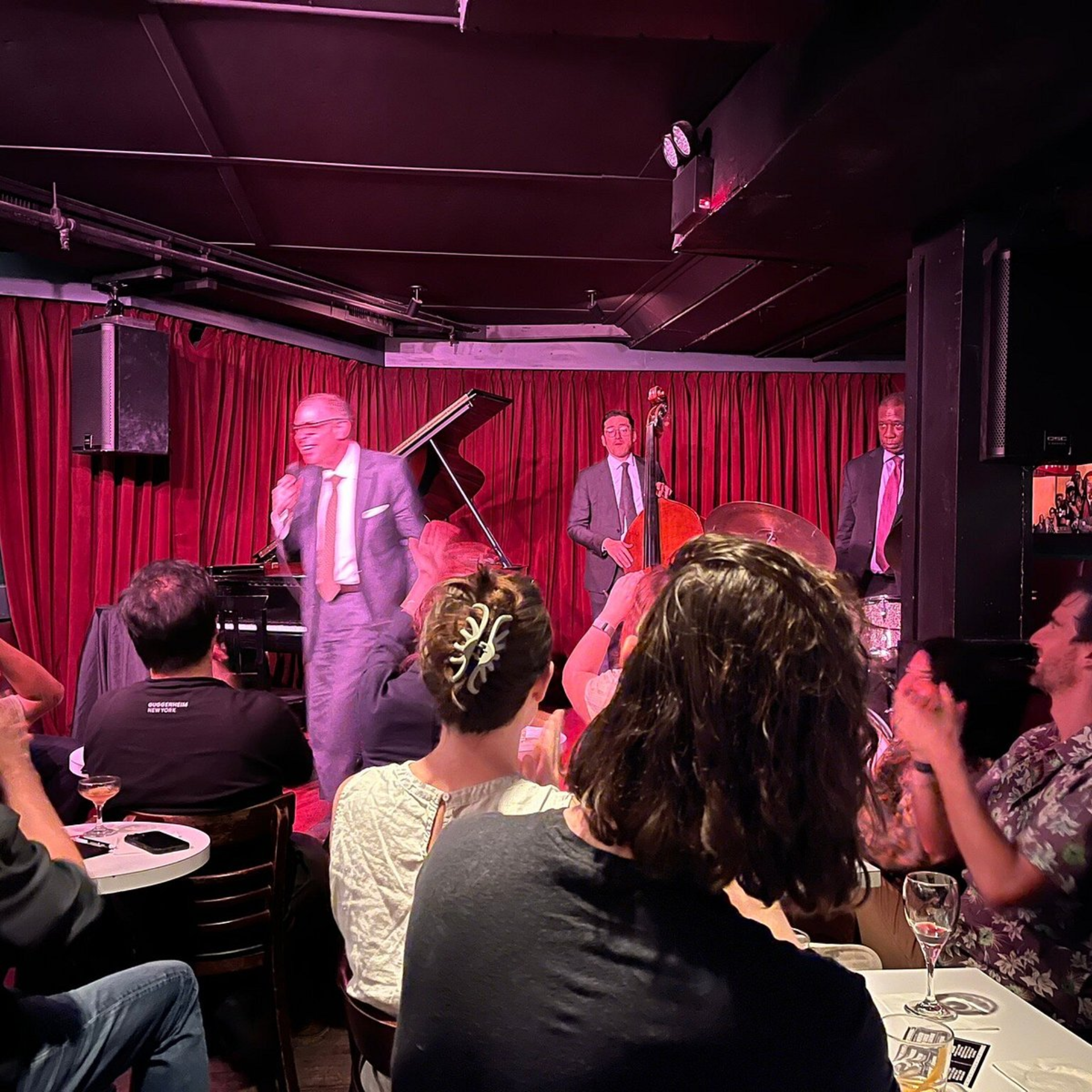 Jazz club performance with singer, upright bass, drums, and piano on stage under red curtains