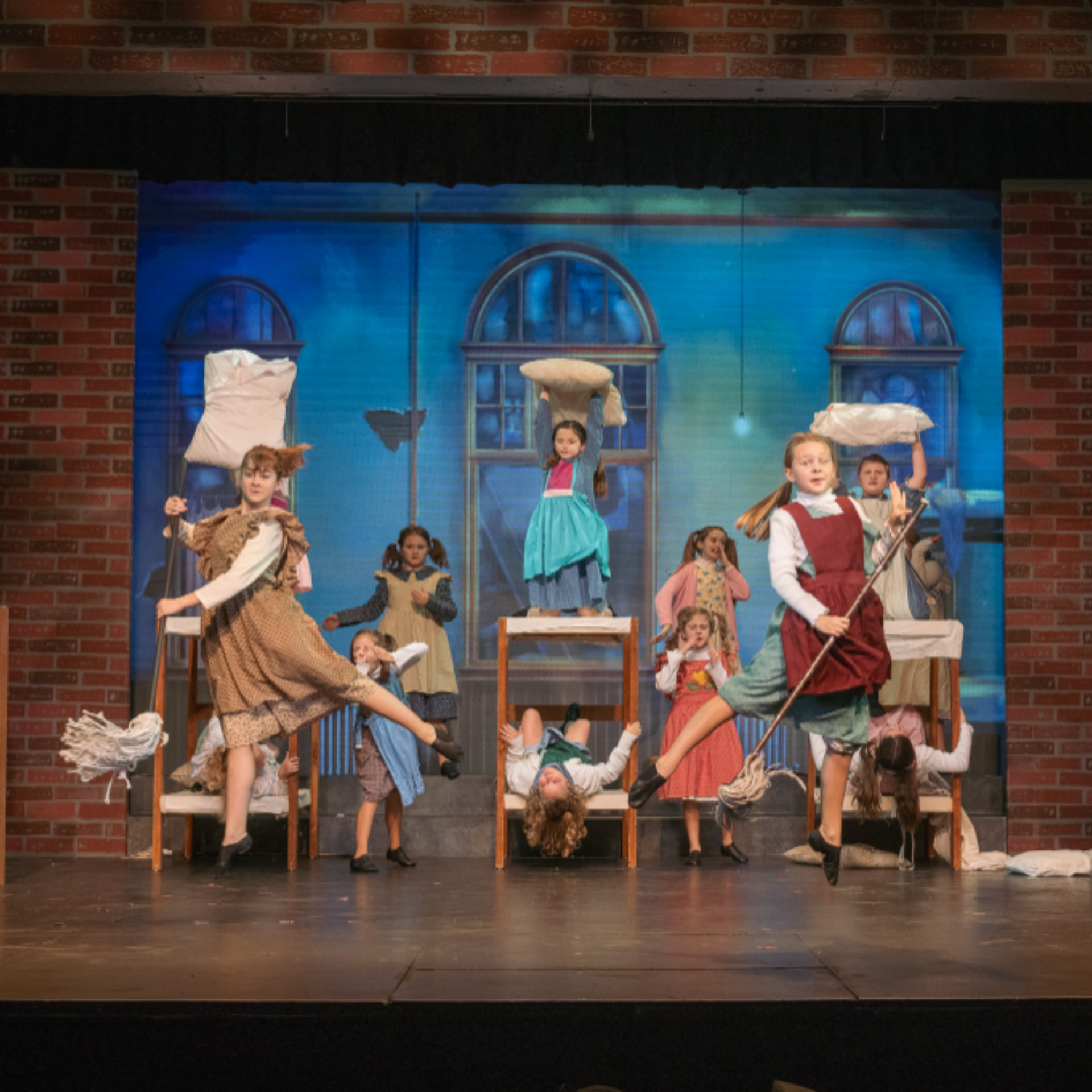 Children performing lively choreographed scene in period costumes on theater stage, using props like mops and brooms, playful orphanage‑style setting with blue‑lit backdrop