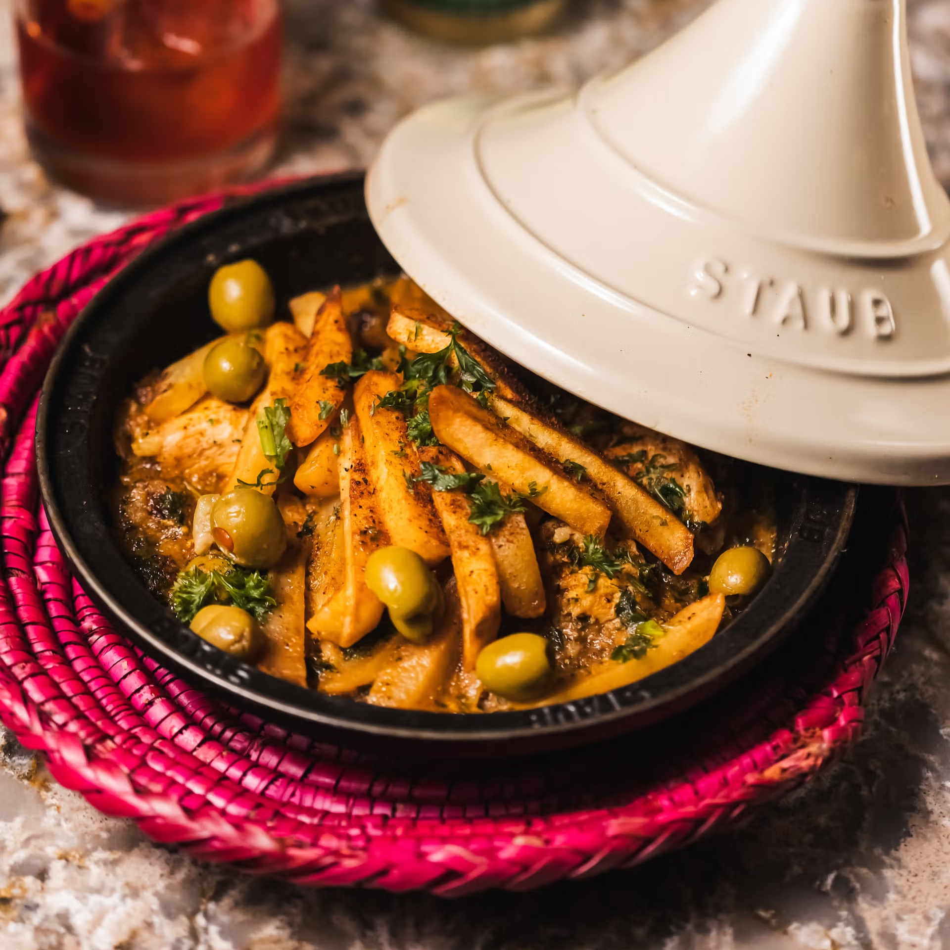 Traditional Moroccan chicken tagine with potatoes, green olives, and herbs served in Staub cookware on woven pink mat, authentic North African cuisine presentation