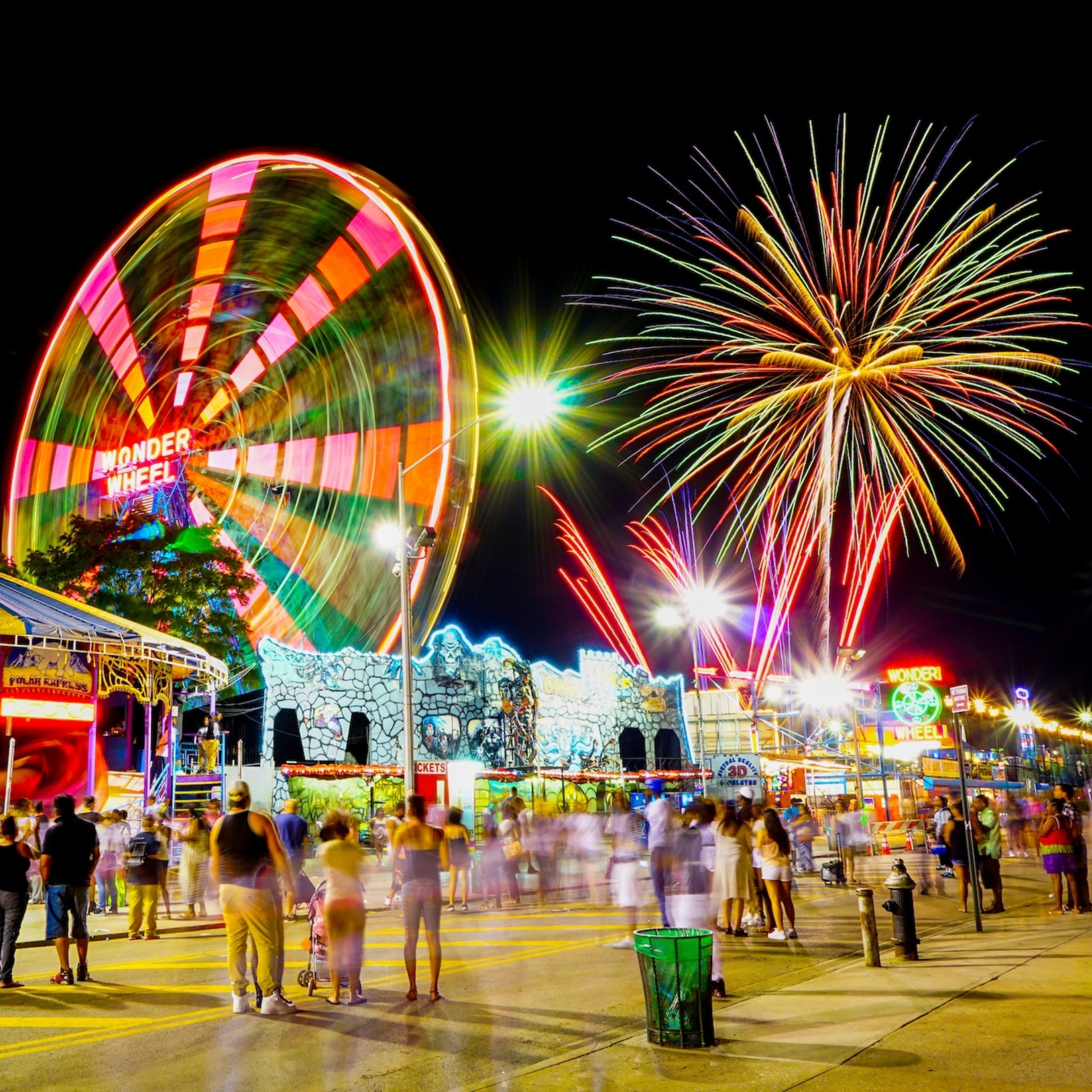 Nighttime amusement park with Wonder Wheel Ferris wheel lit in colorful lights, vibrant fireworks bursting above, neon‑decorated carnival buildings and lively crowd enjoying festive atmosphere
