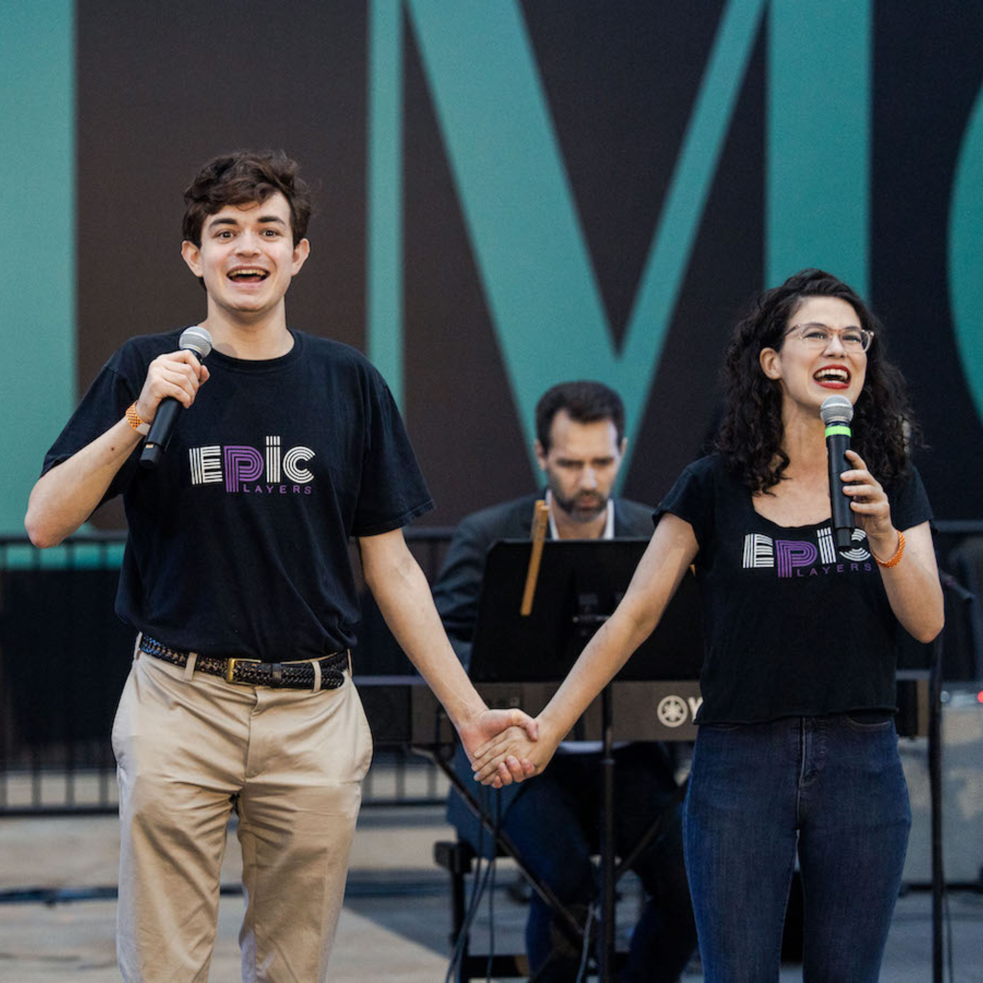 Two performers in black Epic Players shirts holding microphones and hands on stage, accompanied by keyboardist, live inclusive theater performance with teal backdrop