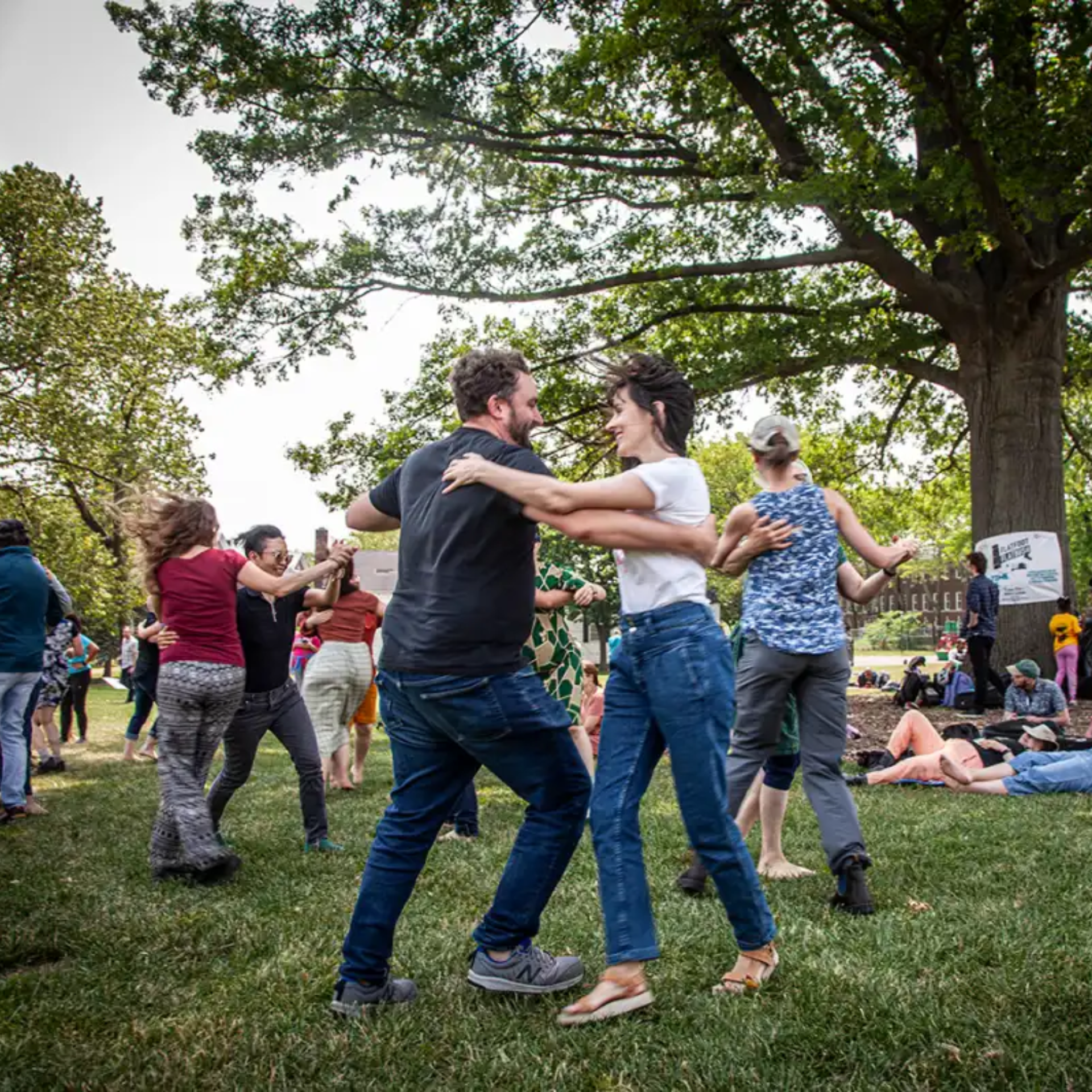 Community outdoor dance gathering on grassy lawn, smiling couples dancing under tall trees, lively social event with people enjoying music and nature