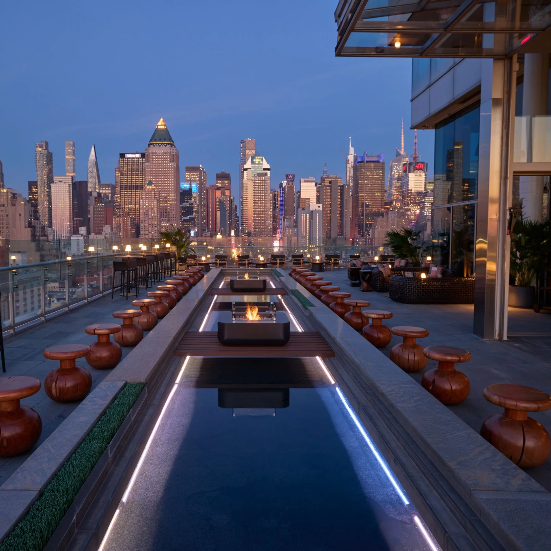 Modern rooftop bar with fire pit water feature and wooden stools overlooking illuminated city skyline at dusk, luxurious lounge atmosphere in New York City