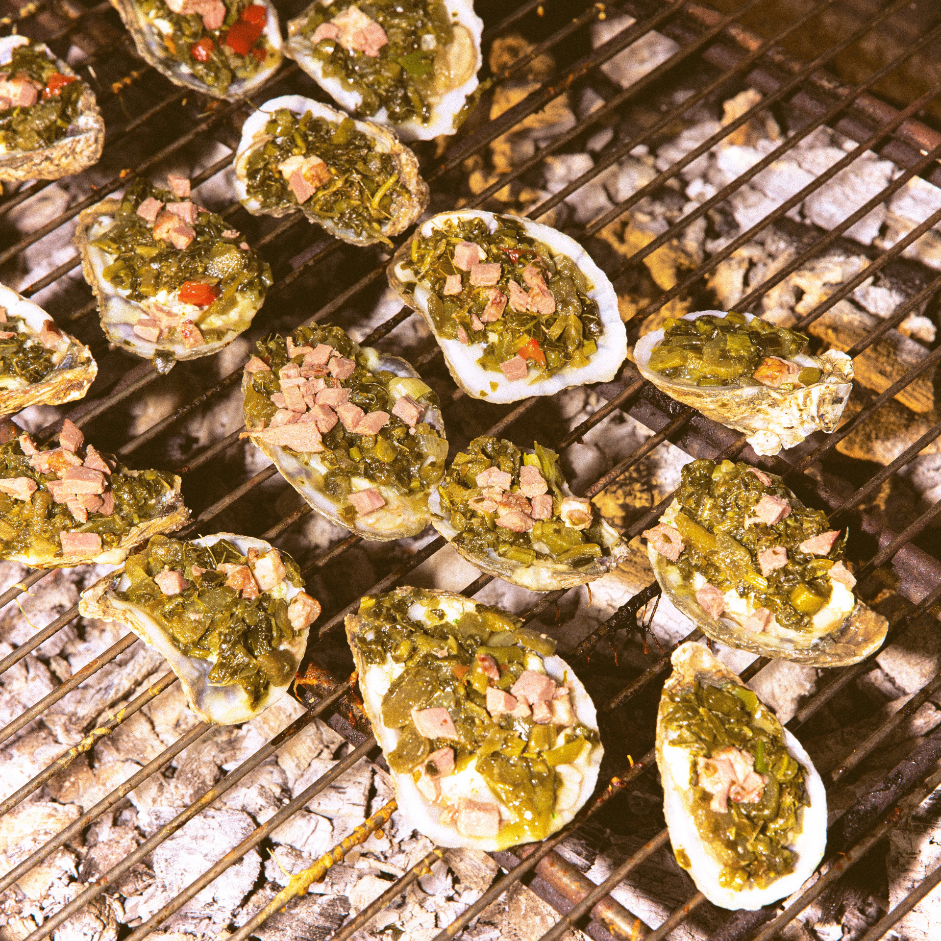 Grilled oysters on a metal grate, topped with herbs and diced meat over glowing coals