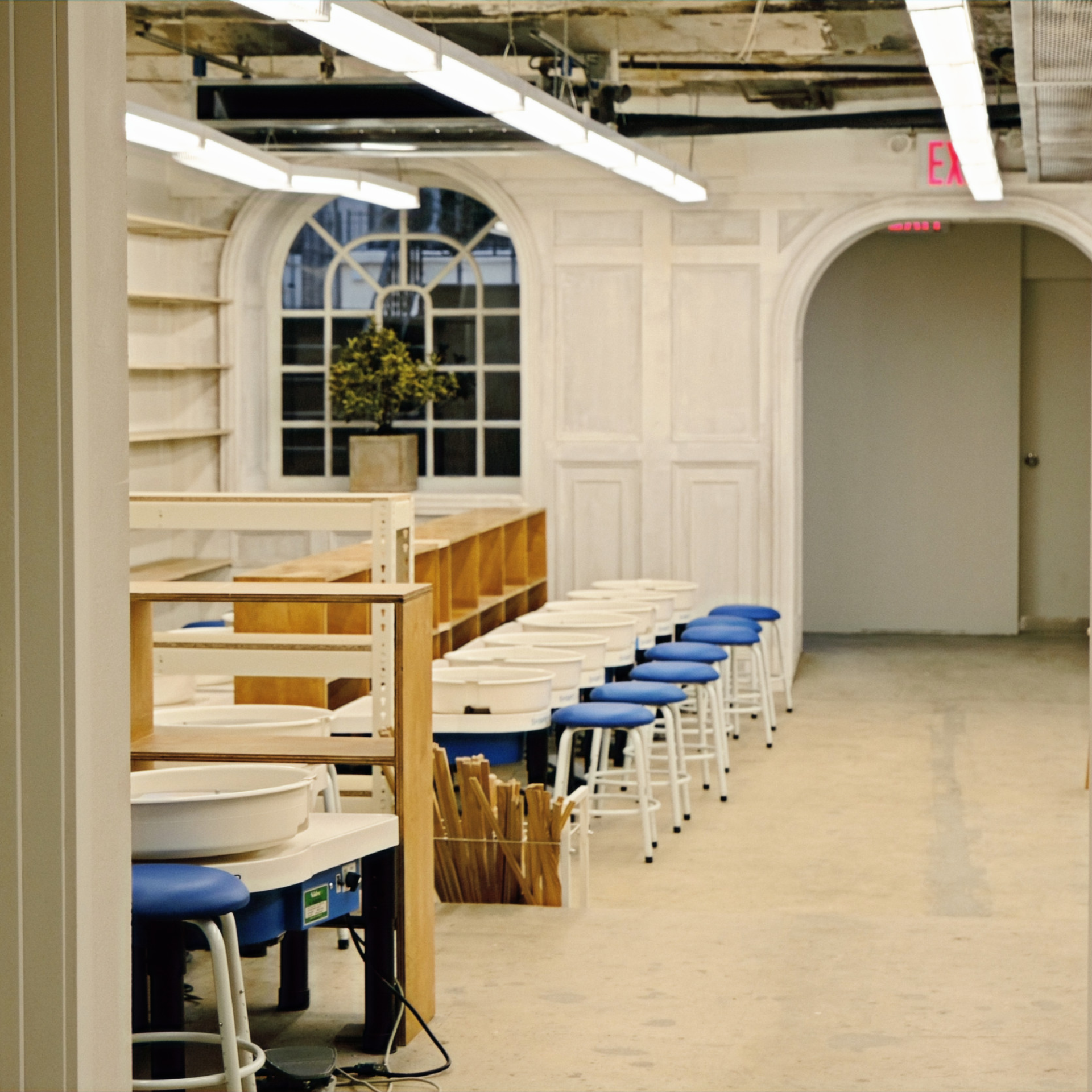 An organized pottery studio featuring a row of white electric pottery wheels paired with blue cushioned stools, set against a backdrop of white wood-paneled walls, an arched doorway, and an arched multi-pane window