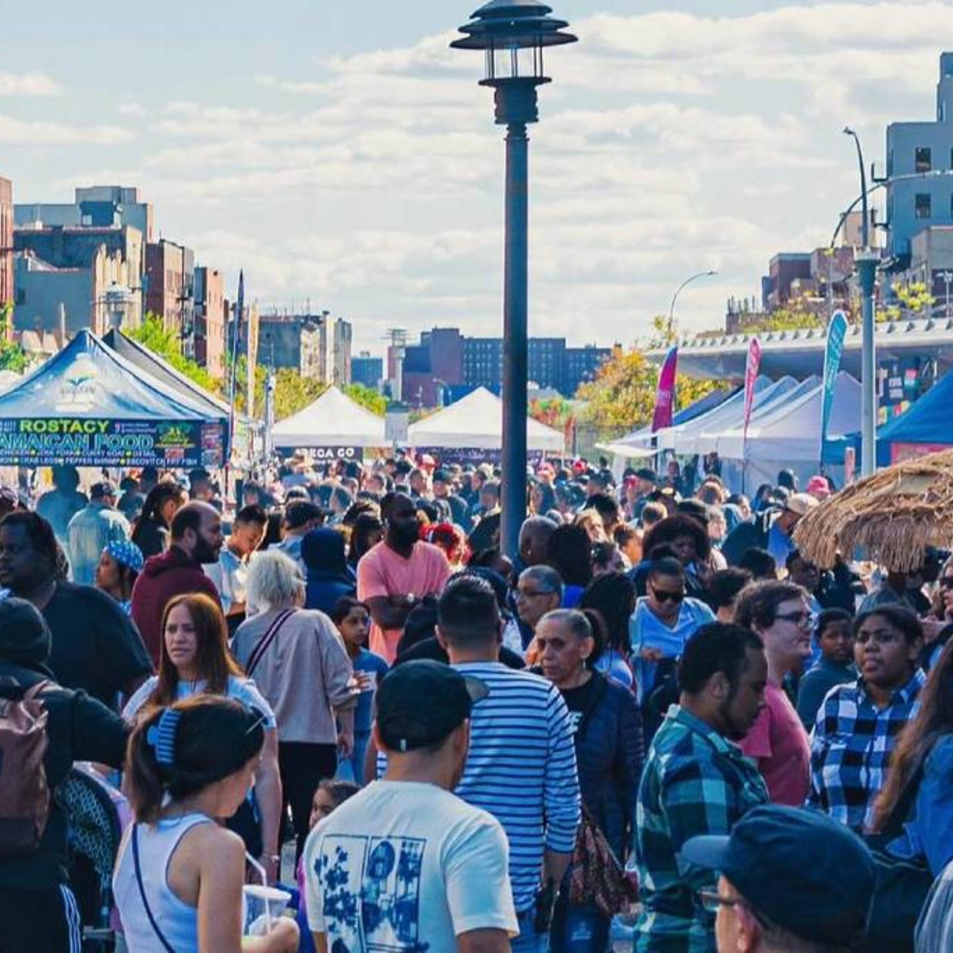 Crowded outdoor street fair with colorful vendor tents, Jamaican food stall, lively crowd walking among local businesses under partly cloudy urban sky