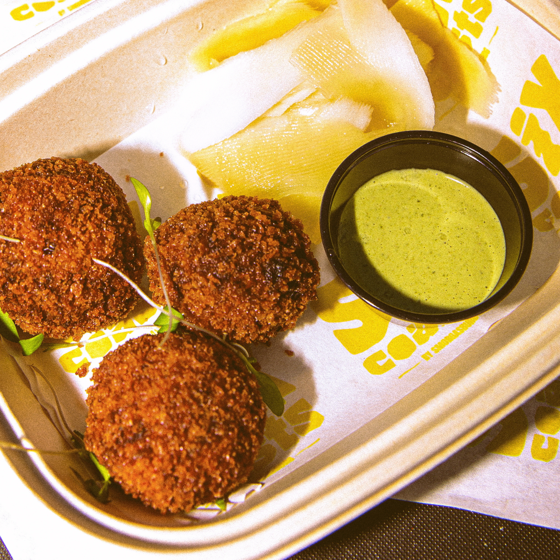 Six Coasts Governors Island: Three golden-brown fried croquettes with green dipping sauce and vegetable slices in a paper-lined container