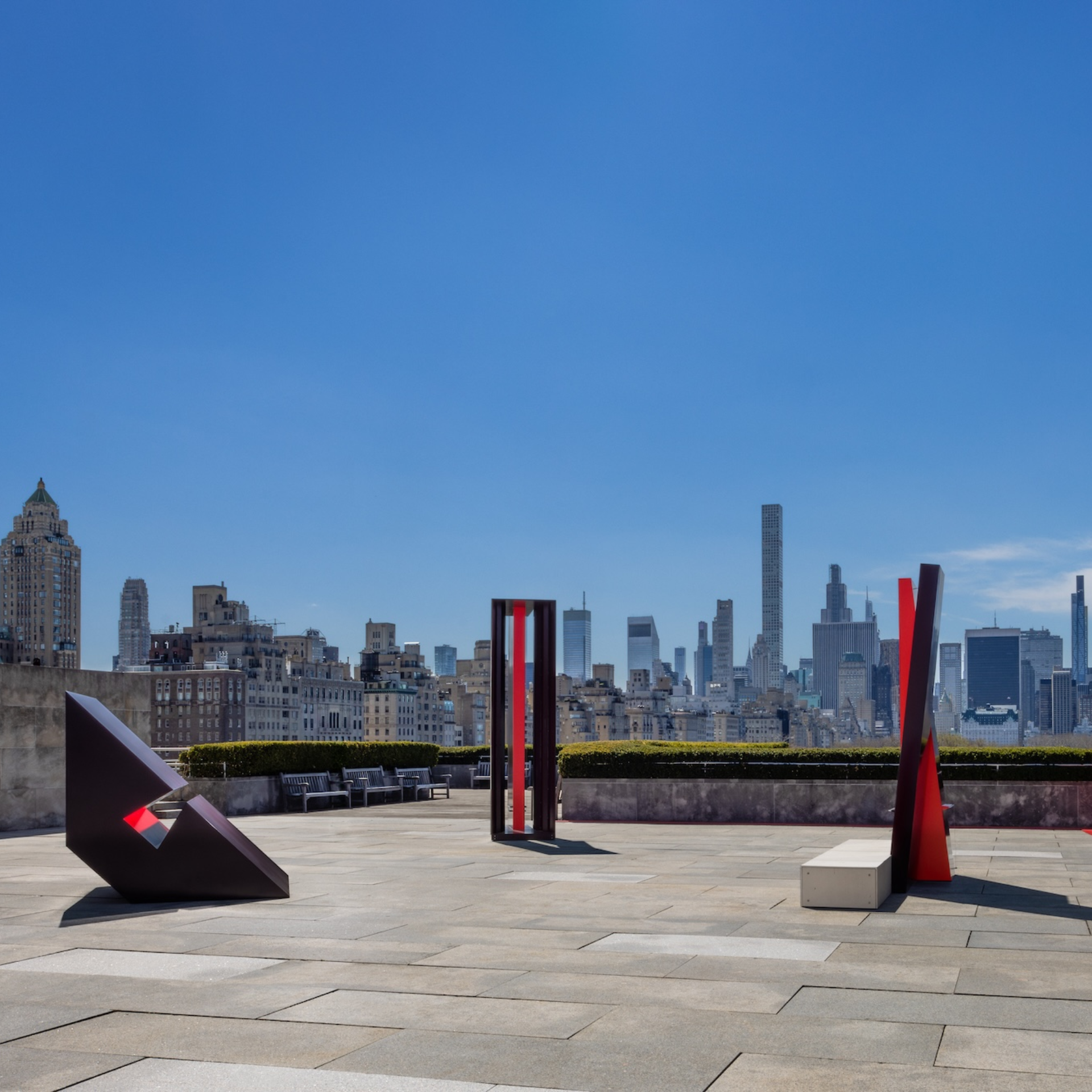 Modern rooftop terrace with abstract red and black geometric sculptures, city skyline of tall skyscrapers in background under clear blue sky, urban art installation blending contemporary design with architecture