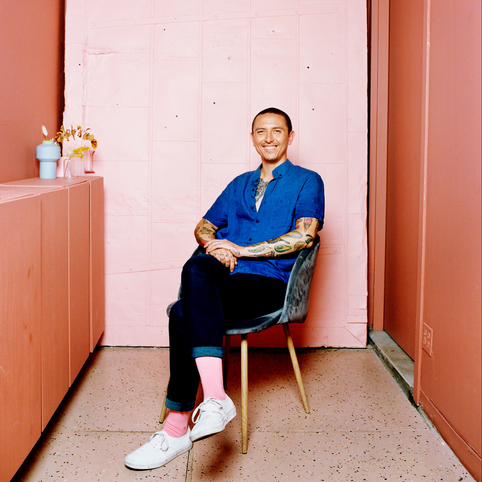 Smiling tattooed person in blue shirt and rolled‑cuff pants sitting in modern pink room with artistic décor, colorful contrast of fashion and interior design