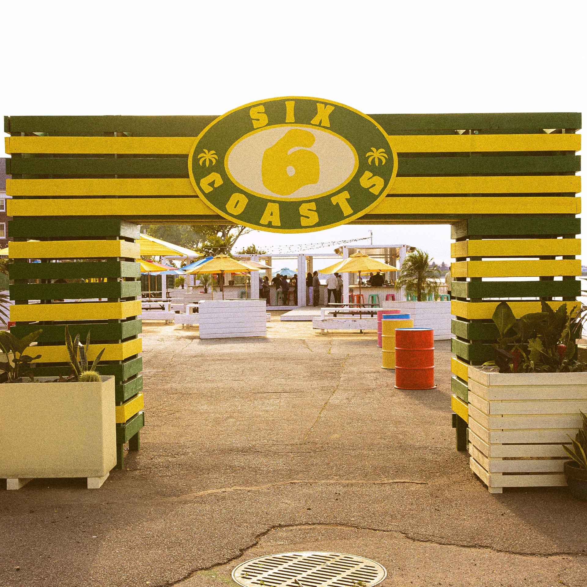 Outdoor venue entrance with green and yellow striped wooden slats, “Six Coasts” sign, and seating under yellow umbrellas