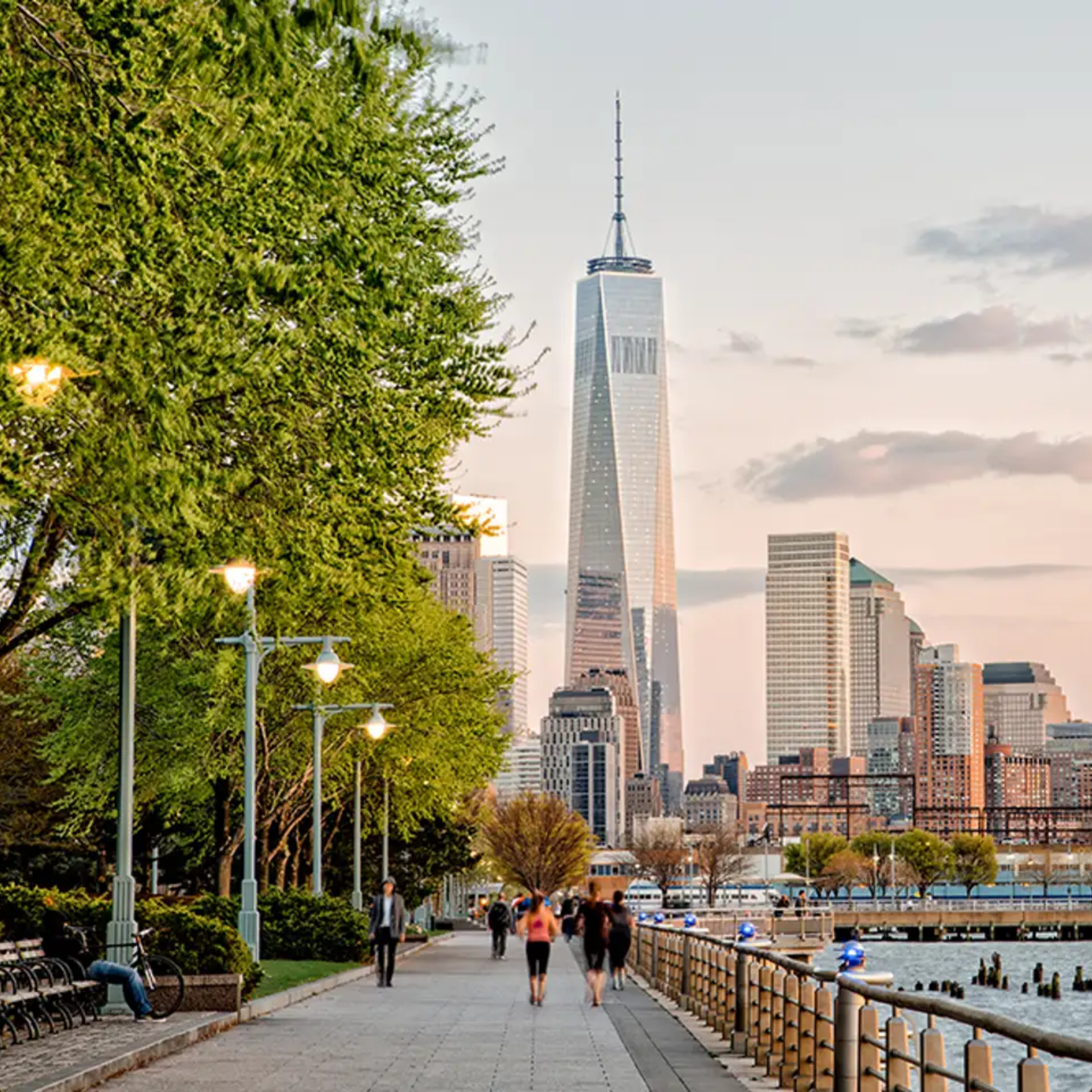 Scenic view of One World Trade Center rising above NYC skyline, waterfront walkway with trees, benches, street lamps, people strolling, and warm sunset tones reflecting on skyscrapers