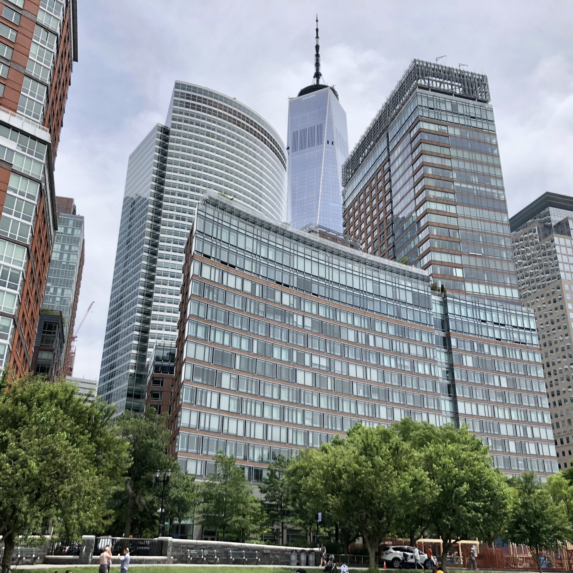 Lower Manhattan cityscape with One World Trade Center spire, modern glass high‑rises, curved and rectangular towers, foreground park with green trees, grass, and people enjoying urban greenery