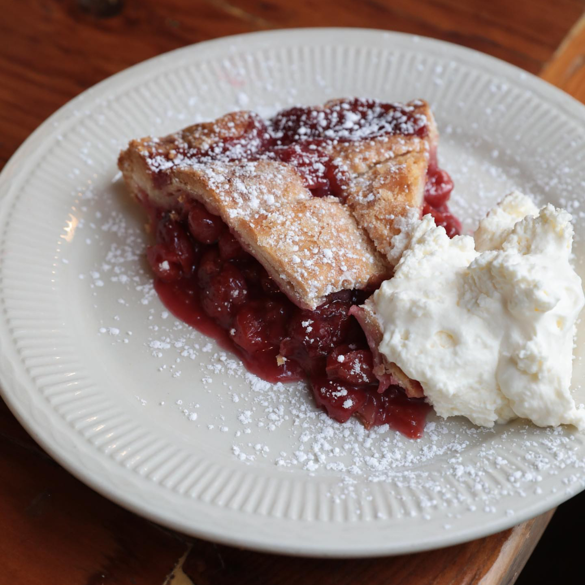 Slice of cherry pie with golden crust, powdered sugar, vibrant cherry filling, whipped cream on white plate, rustic wooden table setting