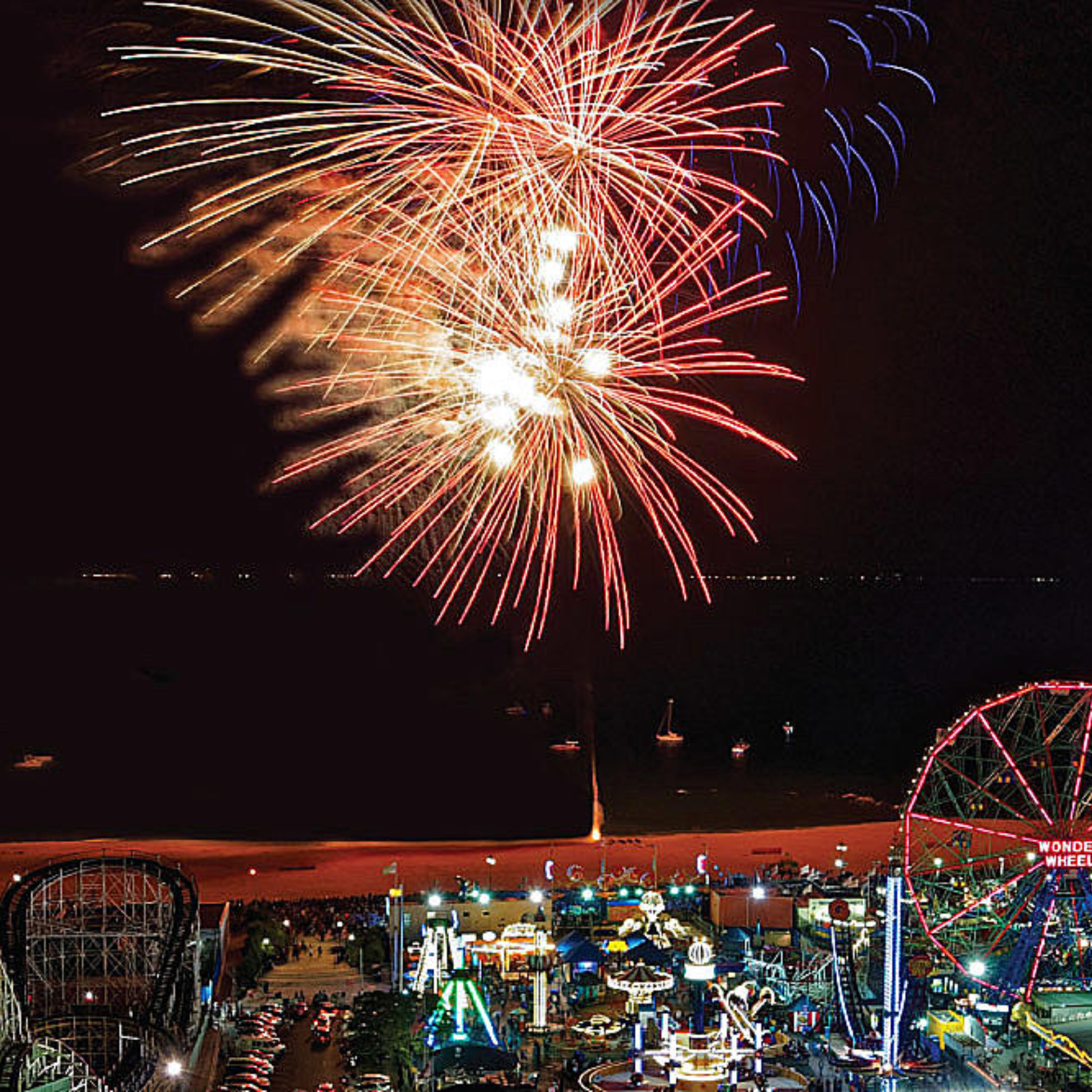 Nighttime amusement park by beach with Wonder Wheel Ferris wheel, roller coaster, glowing rides lit up, colorful fireworks exploding above ocean with boats offshore