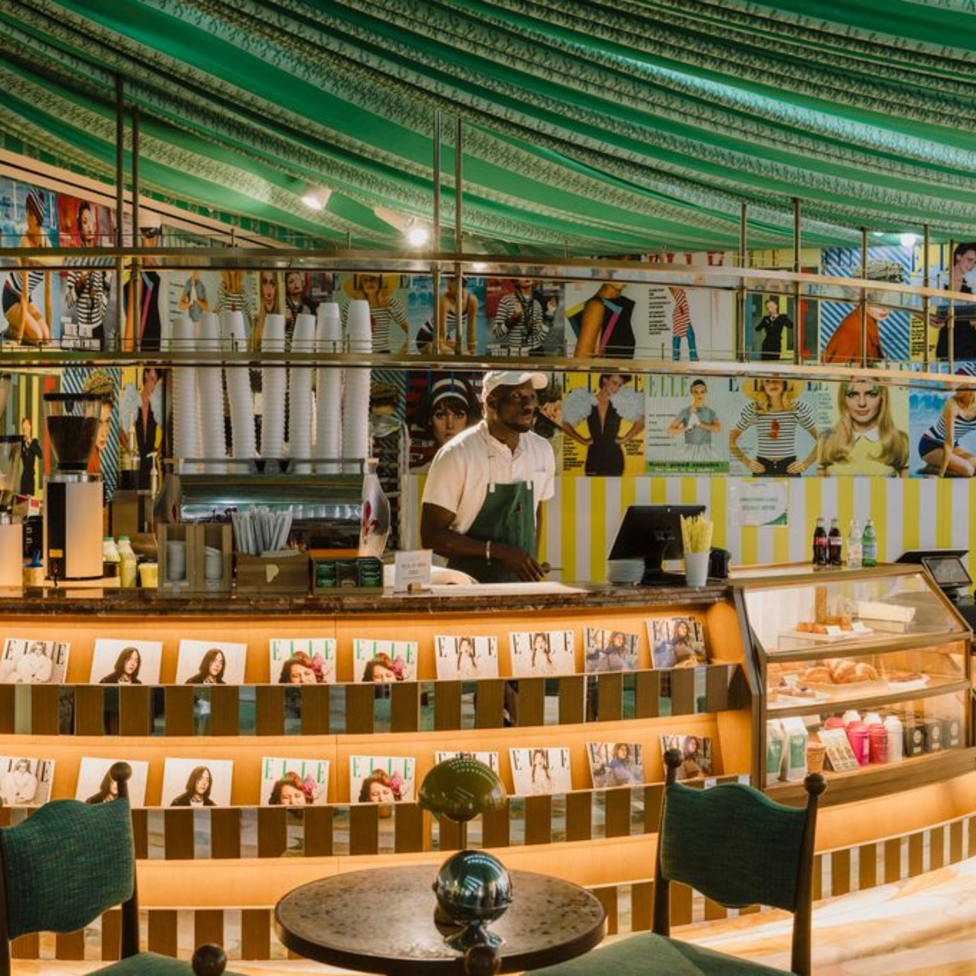 A barista at the counter of a bright, French Riviera-themed ELLE magazine pop-up cafe with yellow and white striped decor and magazine cover displays
