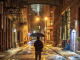 Nighttime urban alley with wet pavement, warm streetlights, person holding umbrella, tall brick buildings with fire escapes, overhead enclosed bridge, and modern towers in background