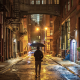Nighttime urban alley with wet pavement, warm streetlights, person holding umbrella, tall brick buildings with fire escapes, overhead enclosed bridge, and modern towers in background