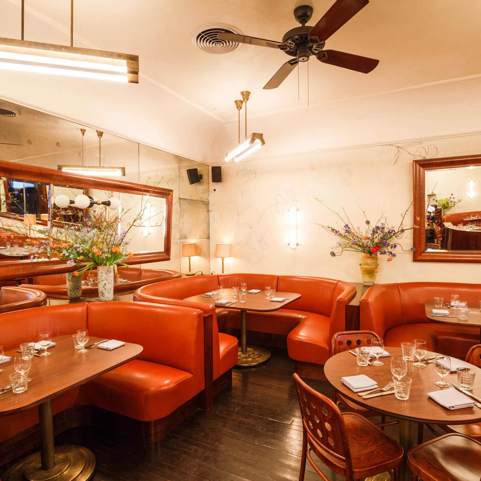 Inviting restaurant interior with curved red‑orange leather booths, dark wooden tables set with glassware, large wall mirrors, warm modern lighting, ceiling fan, and floral arrangements