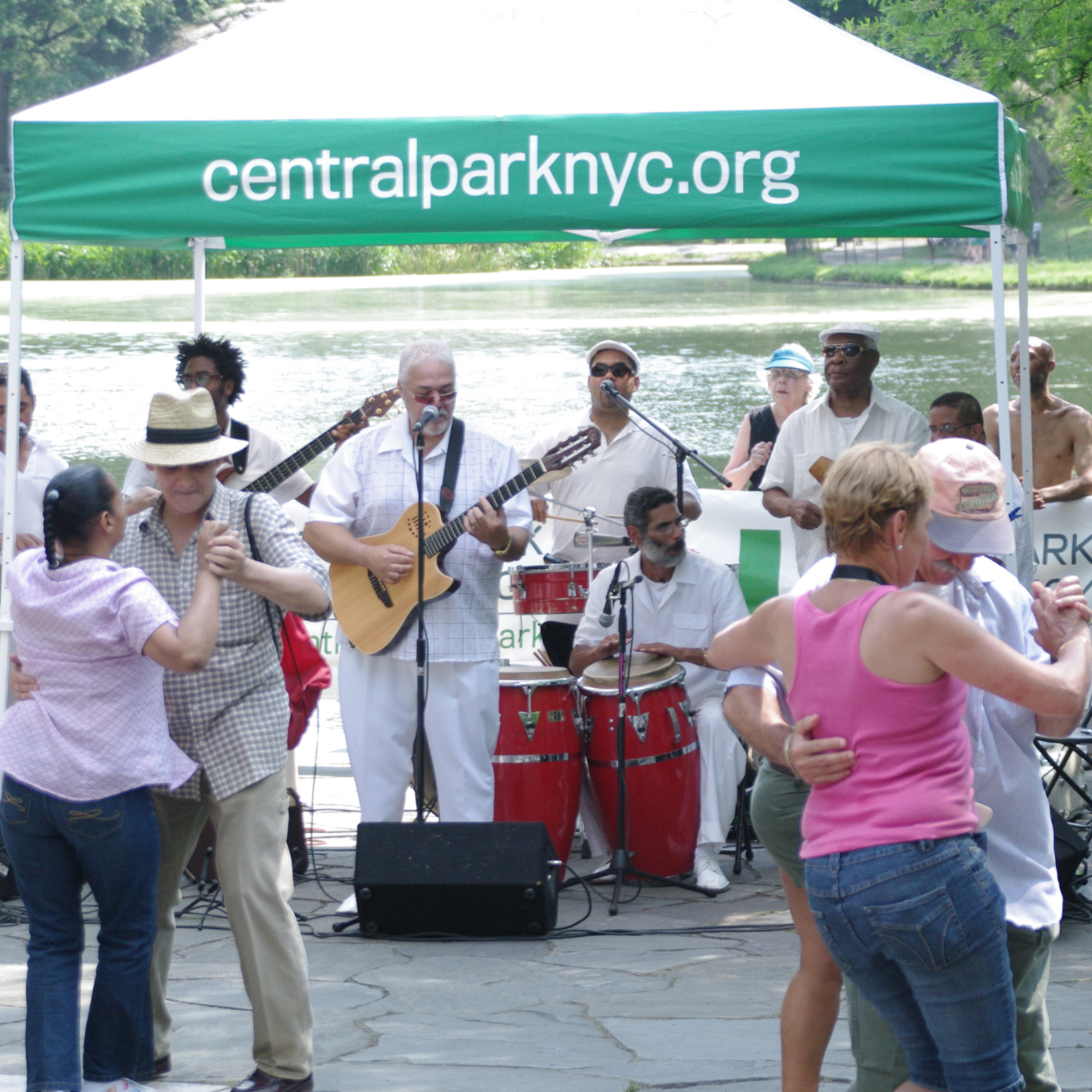 Couples salsa dancing on a stone patio in front of a live Latin music band performing under a green Central Park Conservancy tent next to a lake