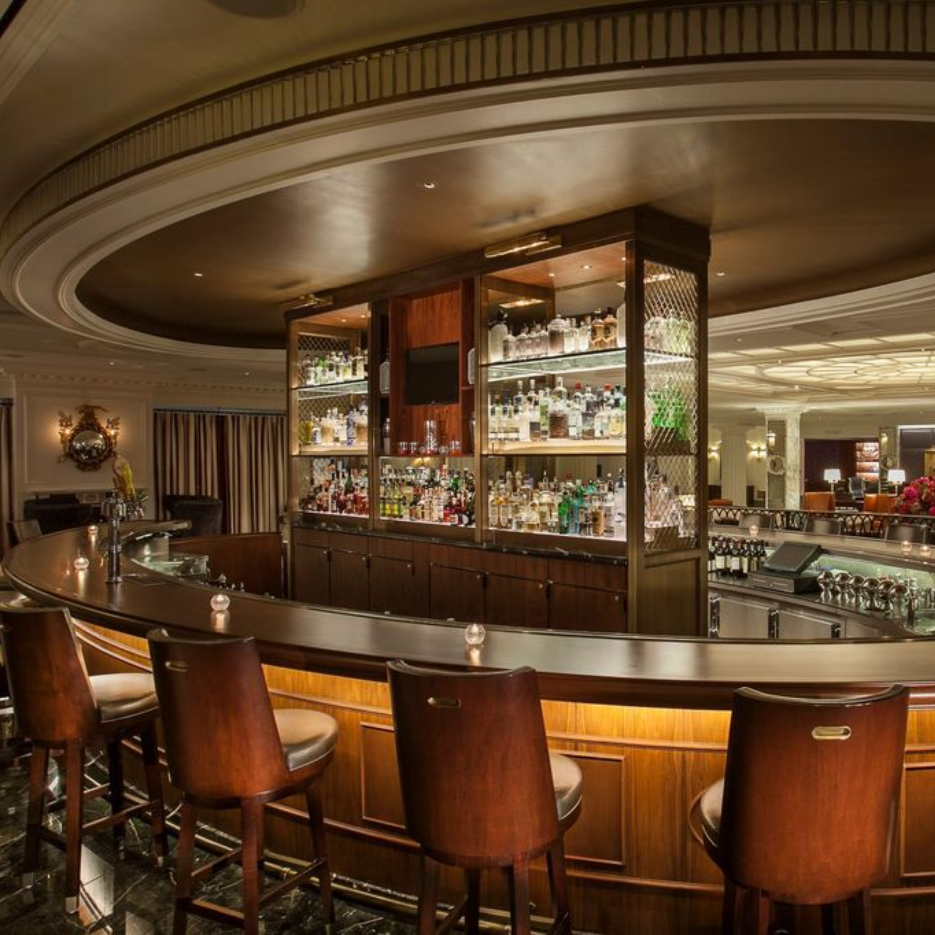 A luxury hotel cocktail bar with a circular wooden counter, leather bar stools, and a glowing, tiered central liquor display shelf