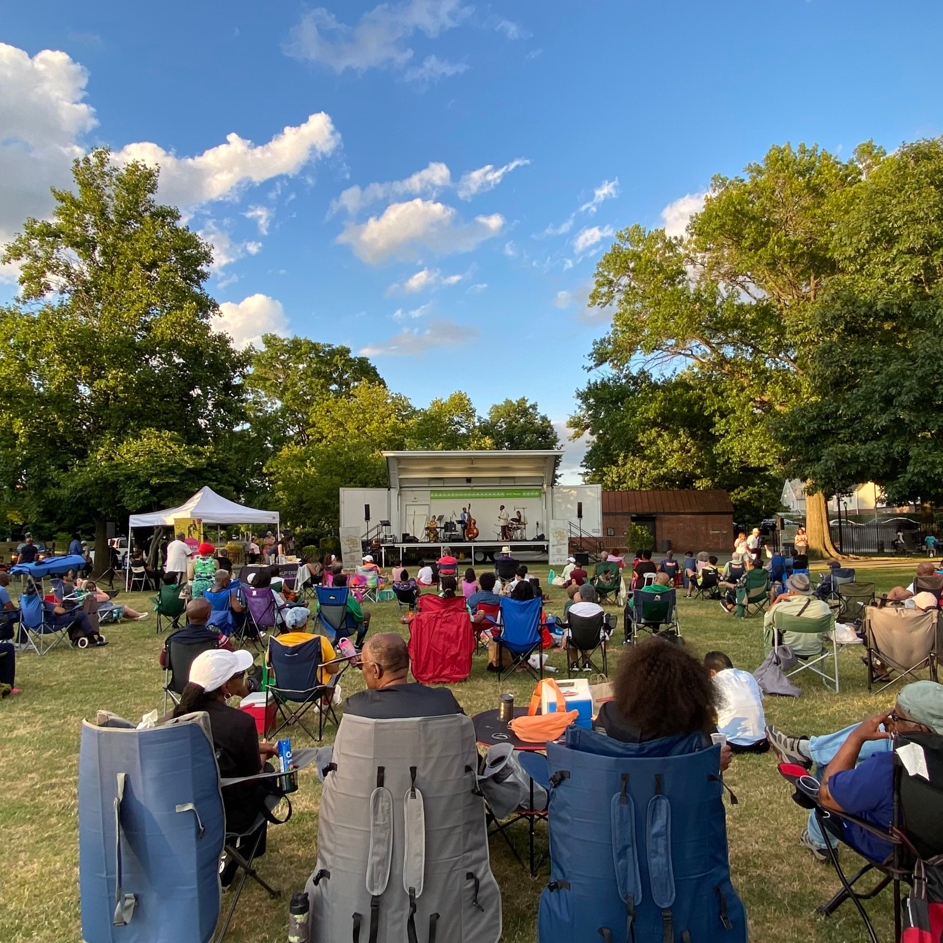 Outdoor park concert with band performing on stage, musicians on guitars and drums, large audience seated on lawn chairs and blankets under trees and blue sky