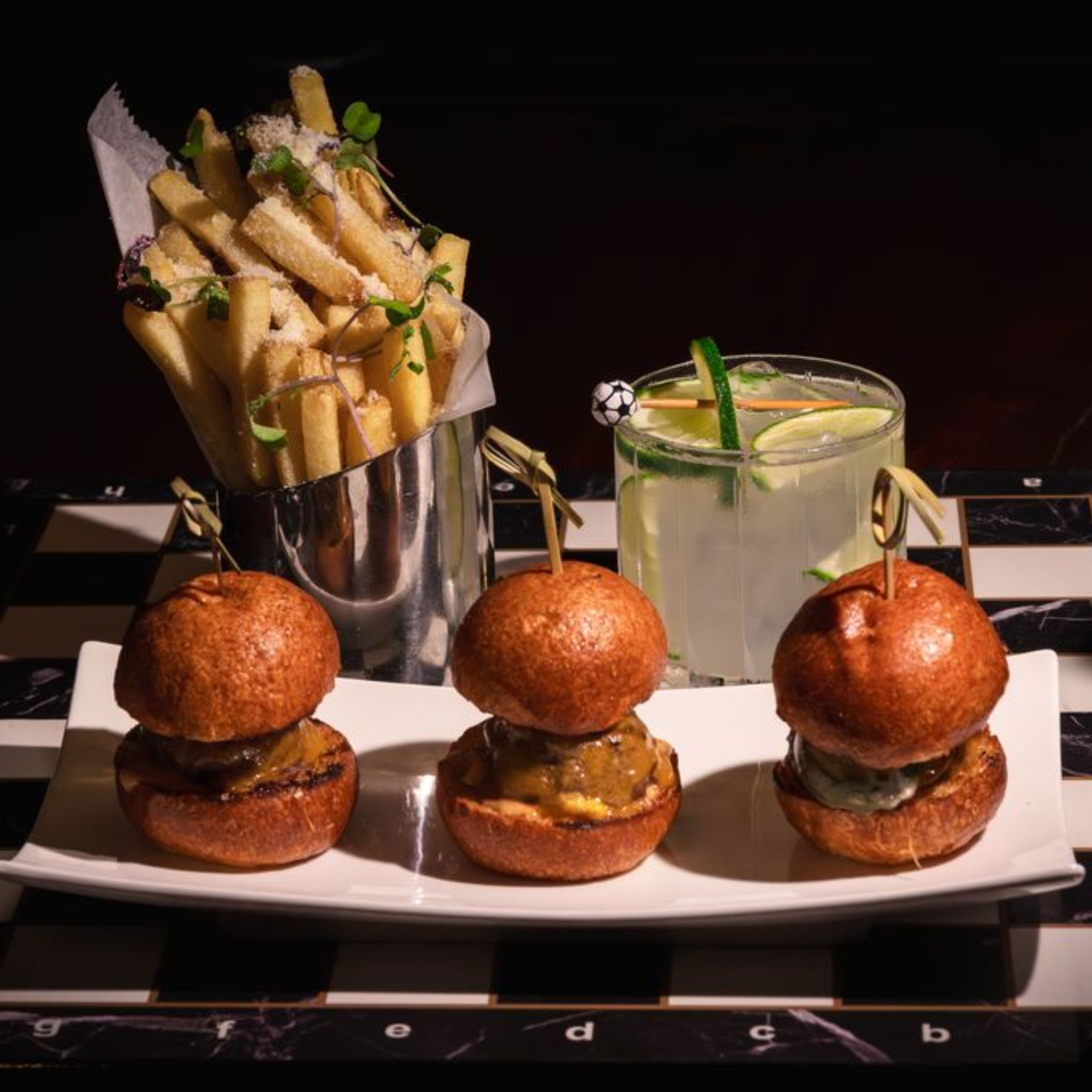 A trio of mini beef sliders, truffle parmesan french fries, and a lime cocktail arranged on a black and white chessboard