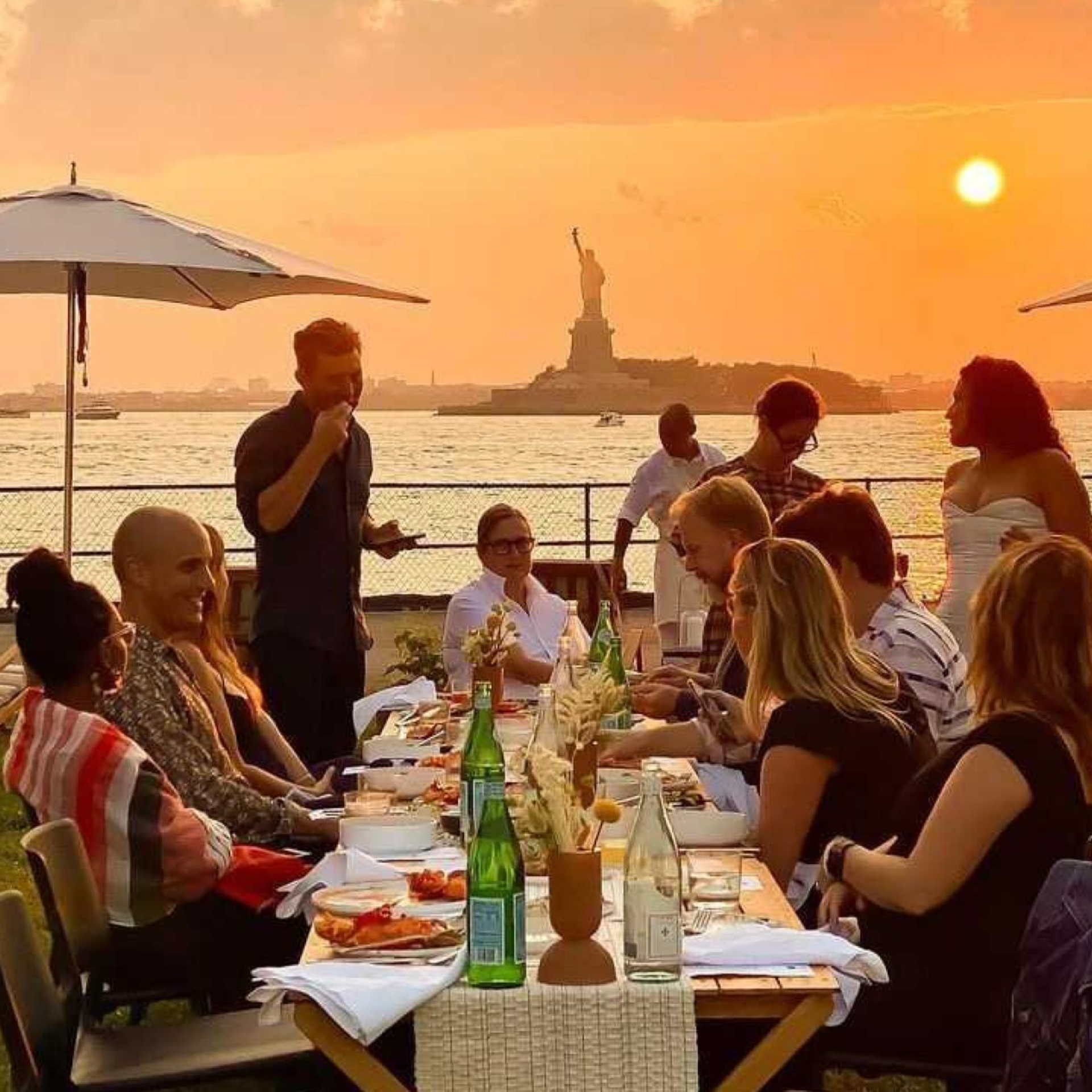 Sunset waterfront dining with Statue of Liberty view, long table gathering, shared plates, boats in background