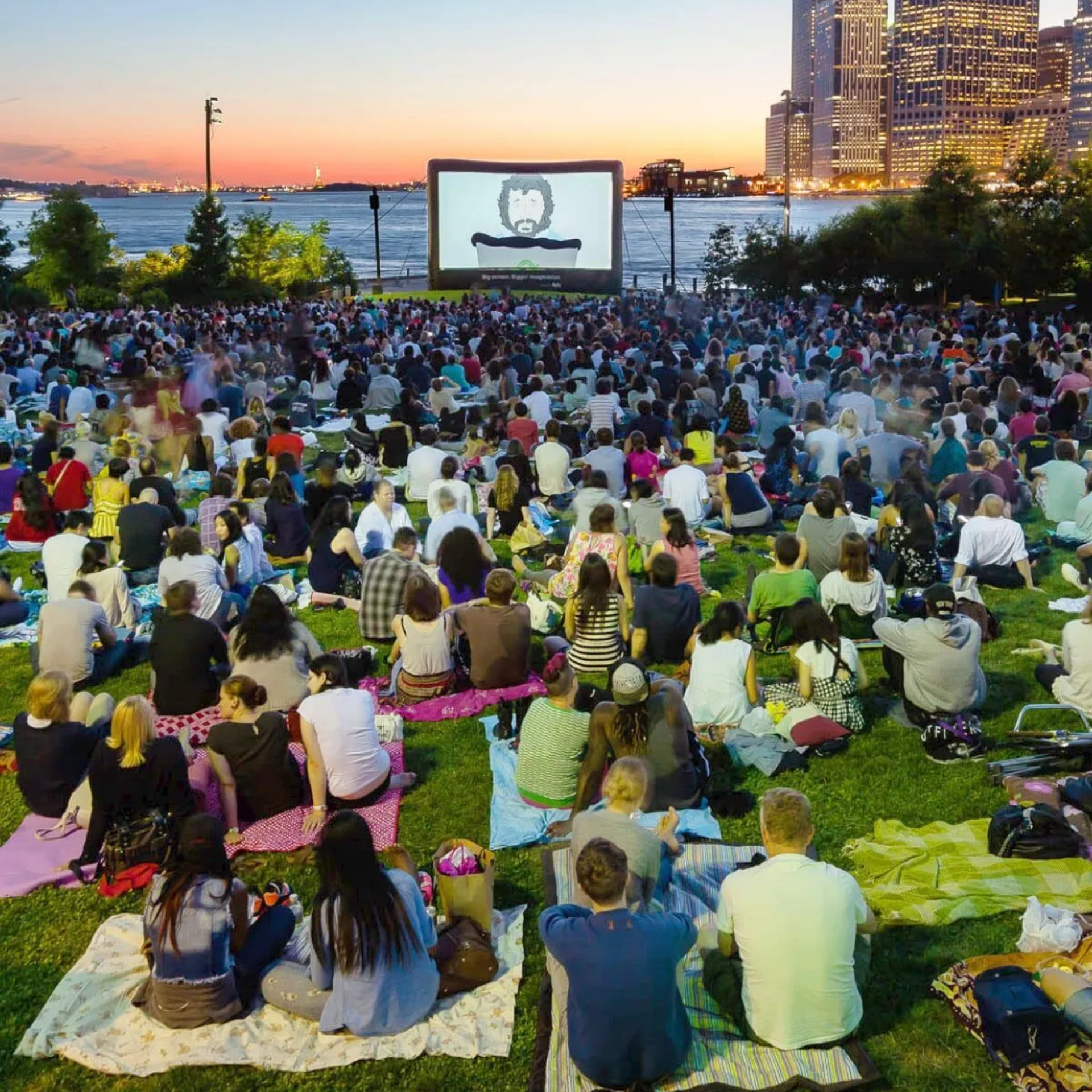 Sunset outdoor movie screening by waterfront with inflatable screen showing illustrated bearded face, crowd seated on blankets and lawn chairs, city skyline and Statue of Liberty in distance
