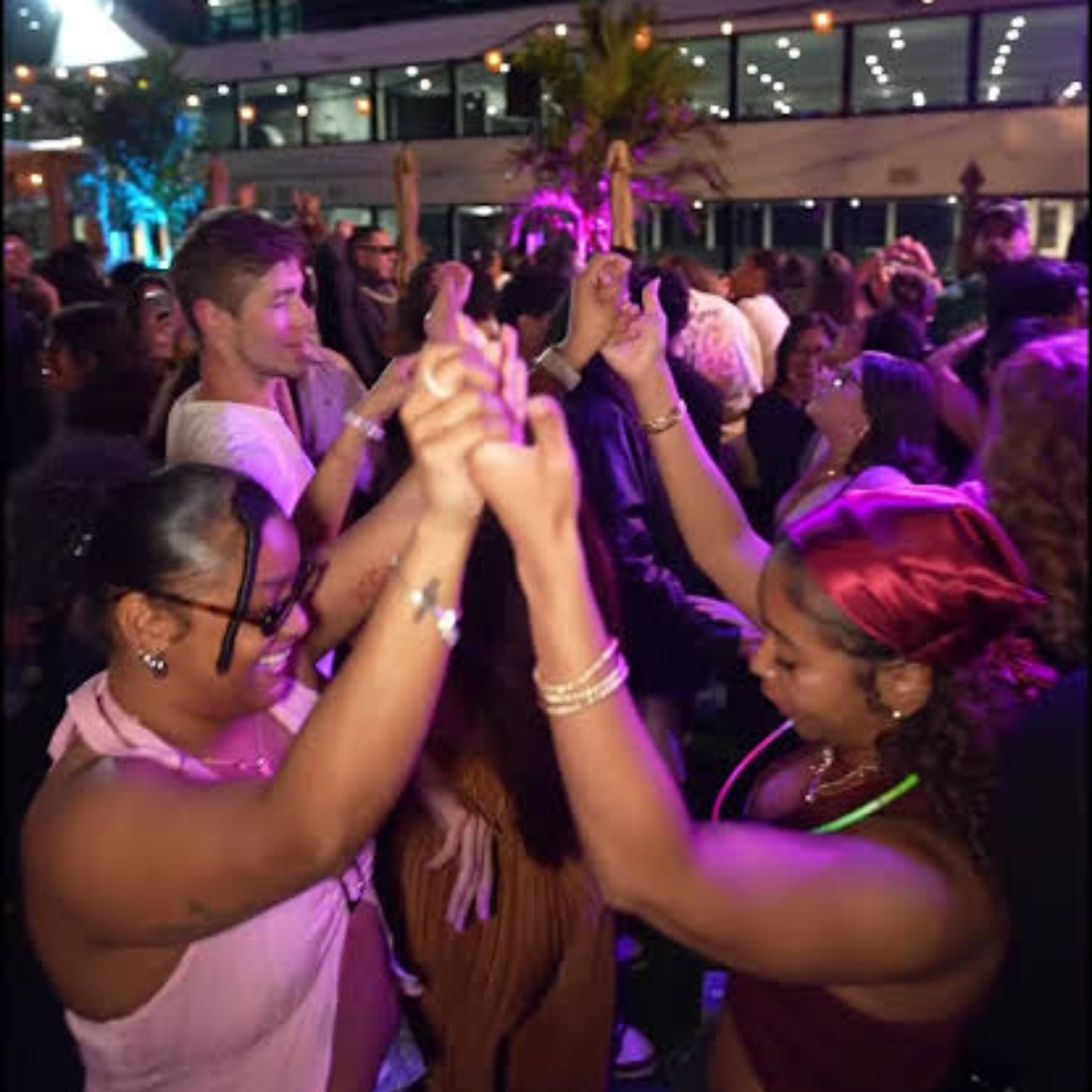 Nighttime outdoor party with crowd dancing, colorful purple and blue lights, palm trees and building in background