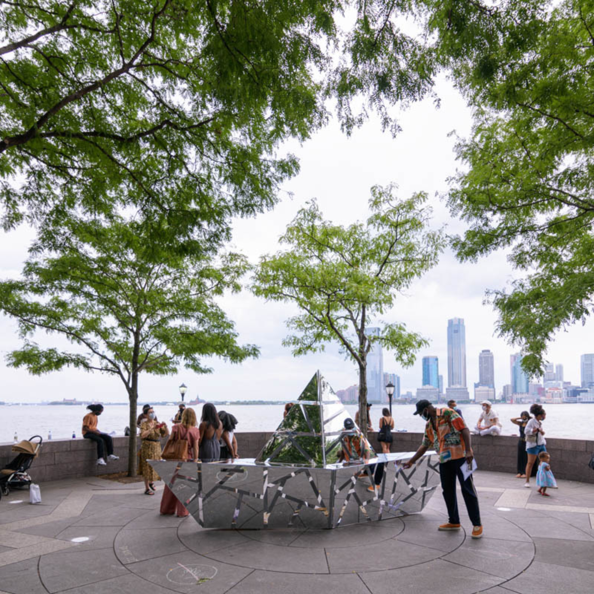 Outdoor public space with mirrored geometric sculpture reflecting trees and people, audience gathered around circular plaza, modern skyscrapers across waterfront skyline
