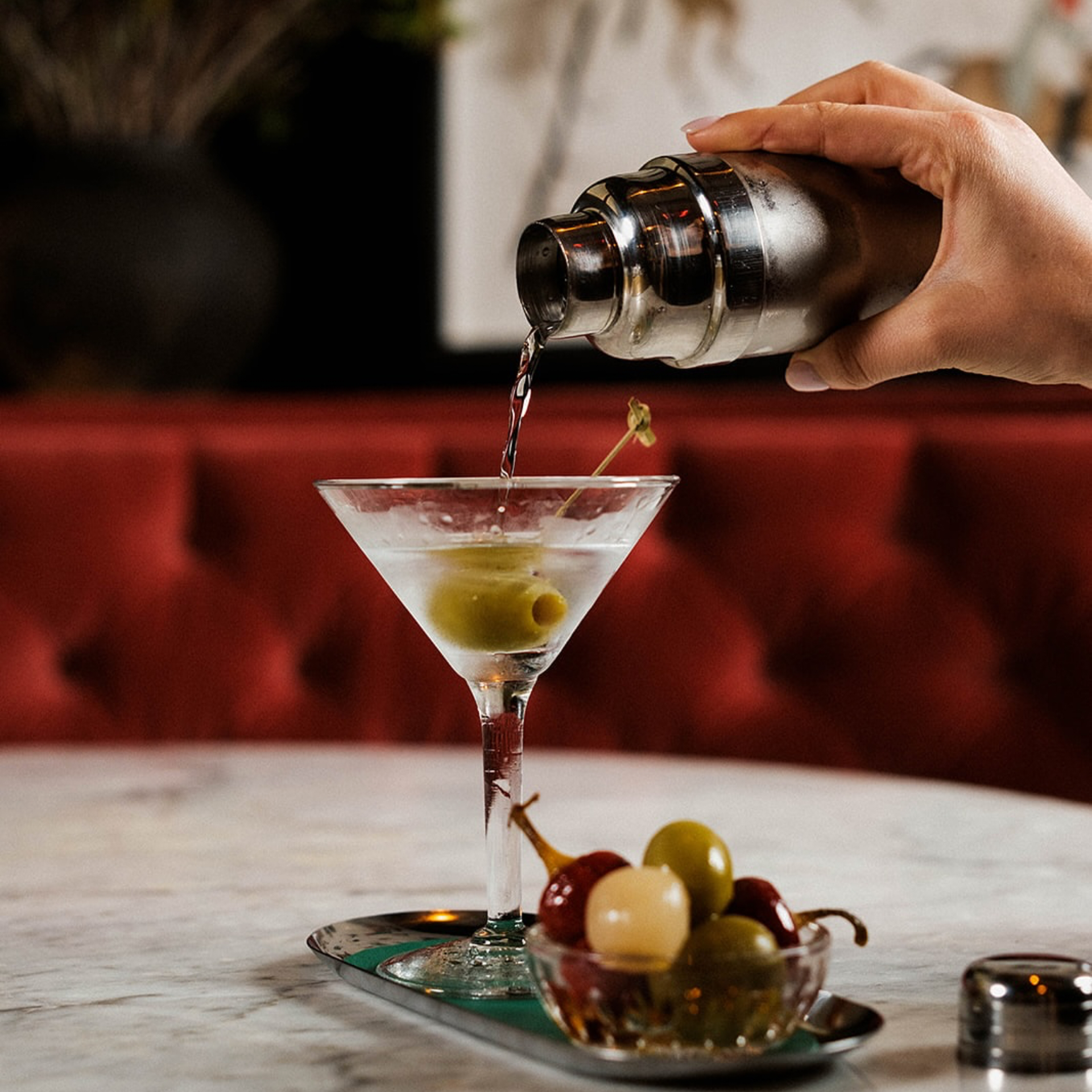 Classic martini poured from stainless shaker into glass with olive garnish, marble table setting, tray of garnishes, and red tufted booth bar background