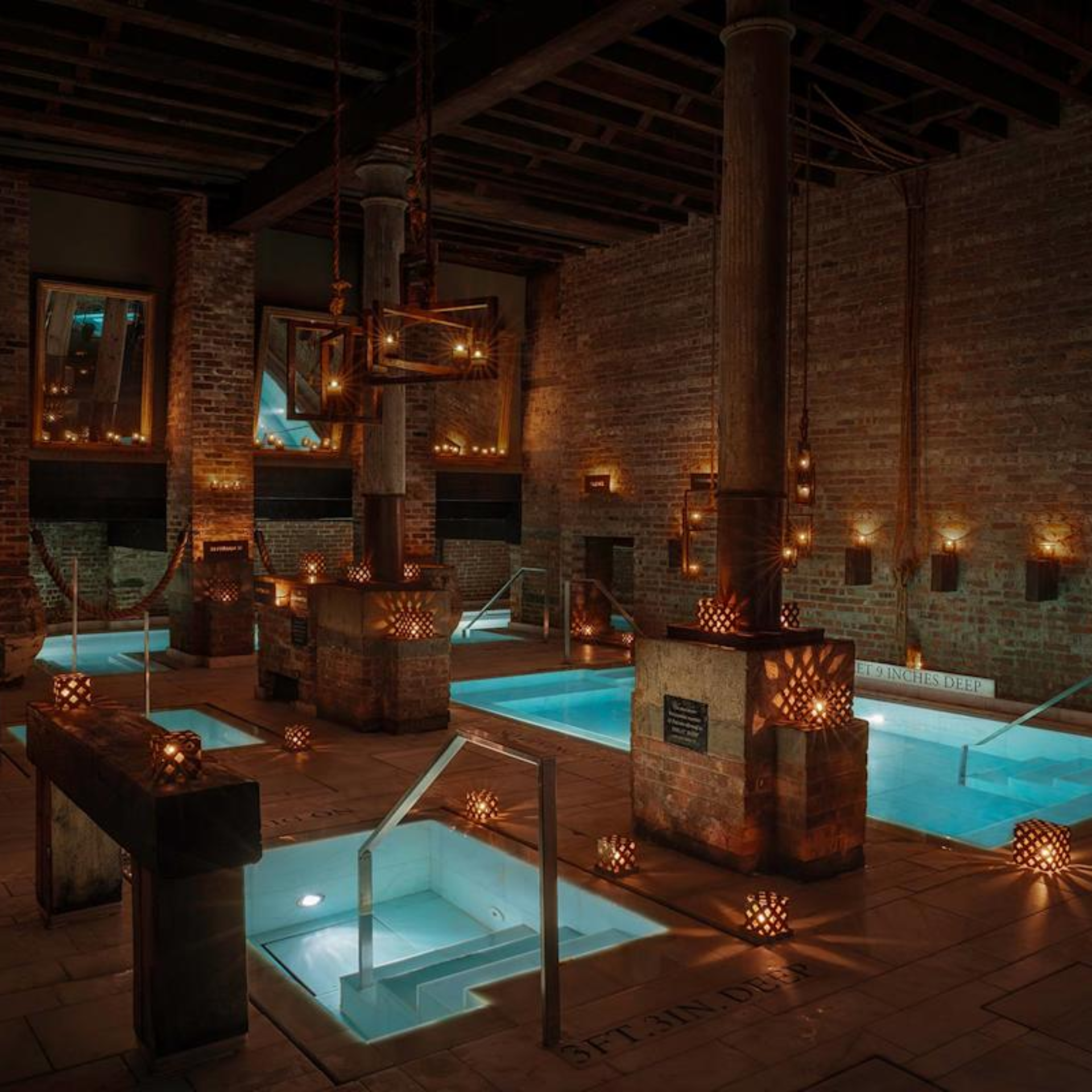 Indoor spa with glowing blue pools, rustic brick walls, wooden beams, and lantern lighting creating a cozy bathhouse ambiance