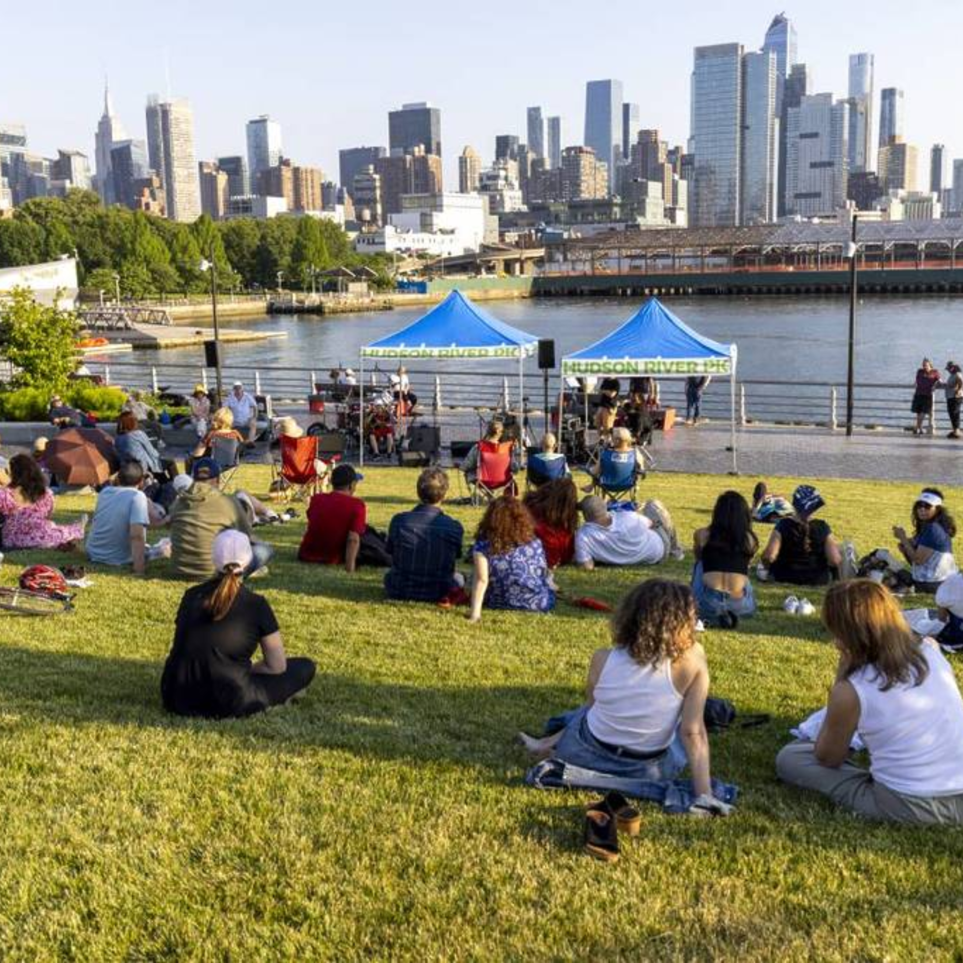 Sunny day community gathering by Hudson River with blue tents, people seated on grass near waterfront, Manhattan skyline of tall modern buildings in background