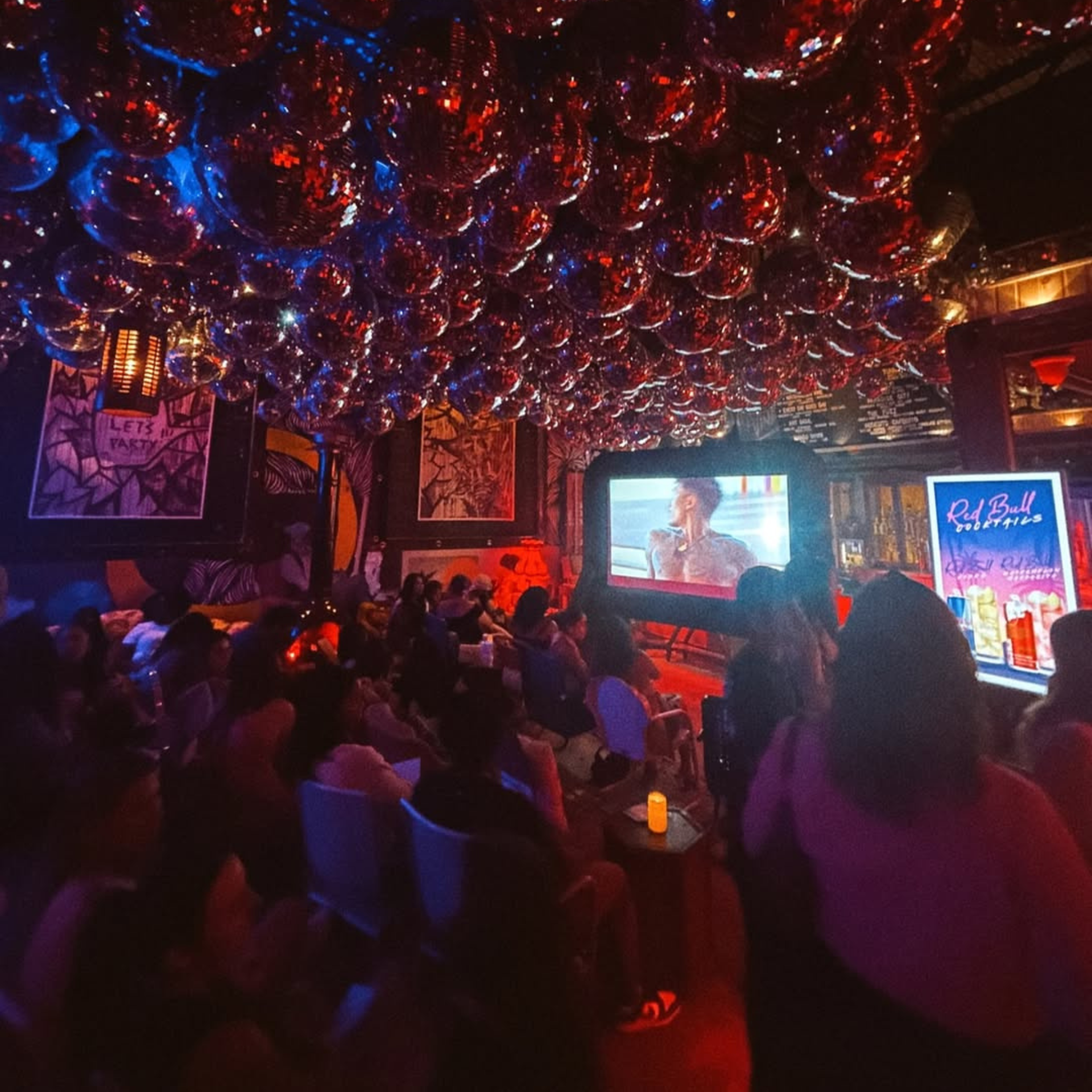 A packed crowd of people at a lounge watch party under a ceiling entirely covered in disco balls, looking at a large television projection screen