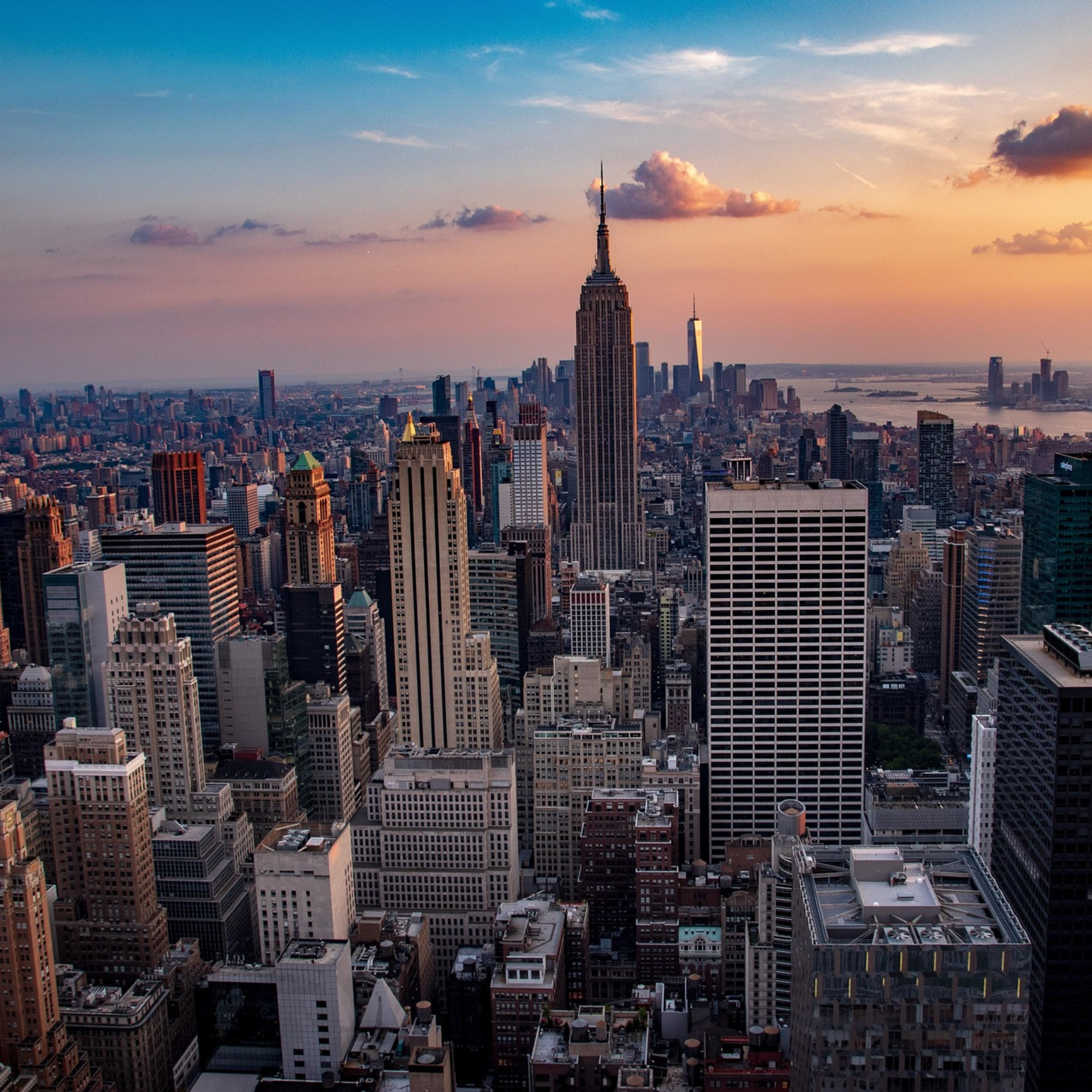 Panoramic New York City skyline at sunset with Empire State Building, One World Trade Center, Hudson River, and skyscrapers under orange‑pink clouds