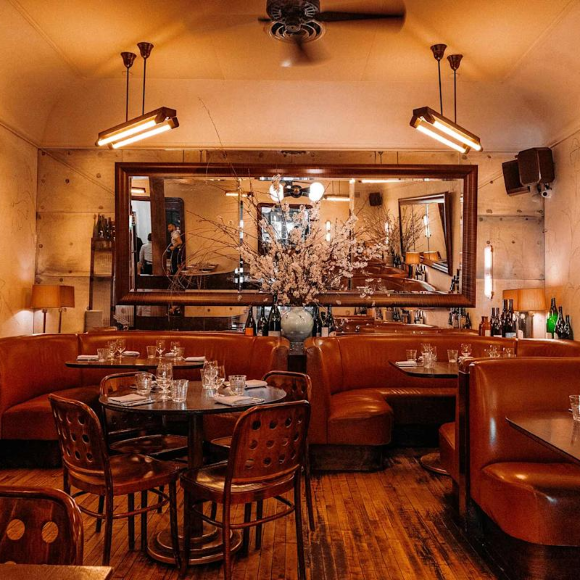 Cozy vintage‑style restaurant with brown leather booths, dark wooden tables set with glassware and napkins, warm amber lighting, decorative vase with blossoms, large wall mirror, and inviting elegant atmosphere