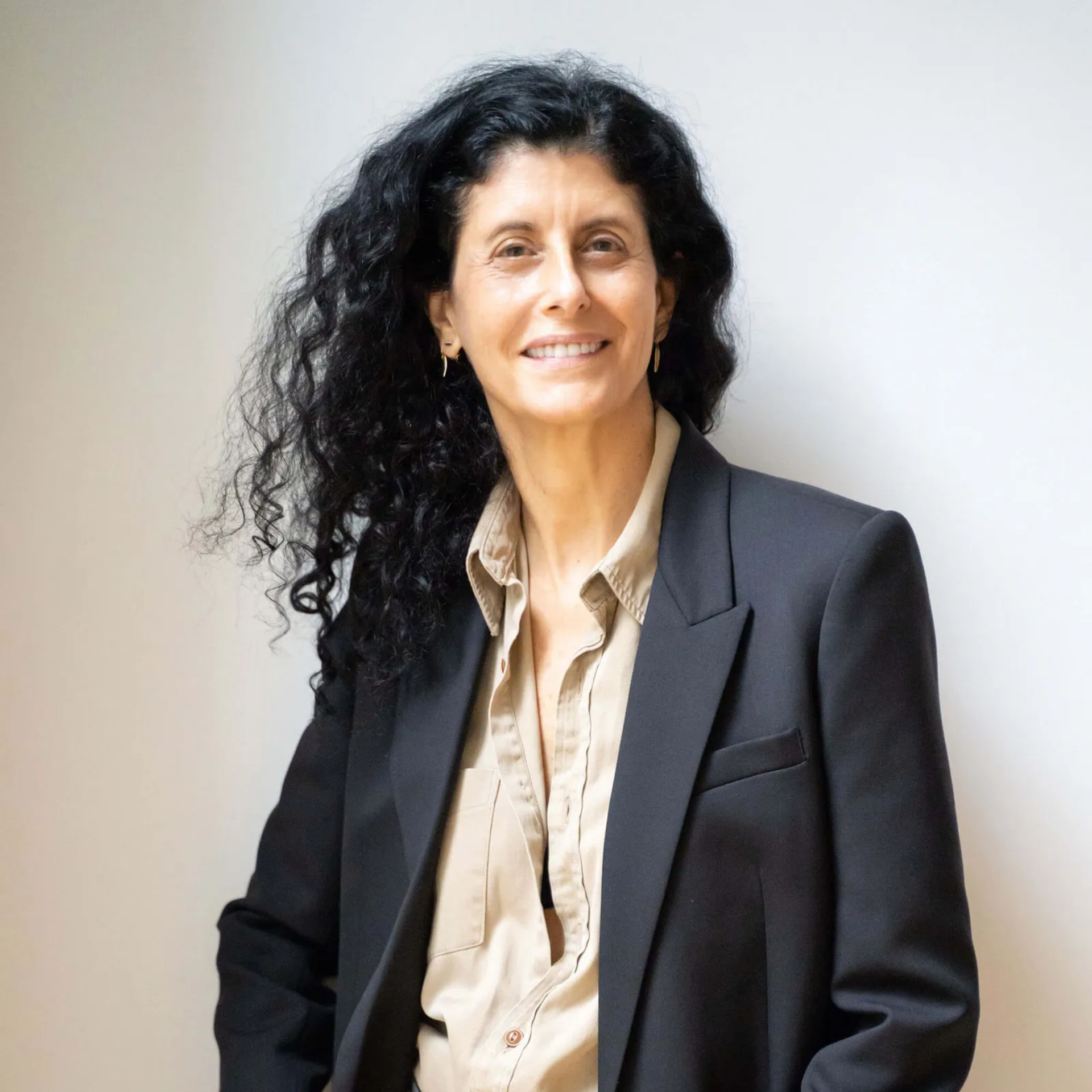 Professional portrait of person with long curly dark hair wearing beige button‑up shirt and black blazer against plain light background