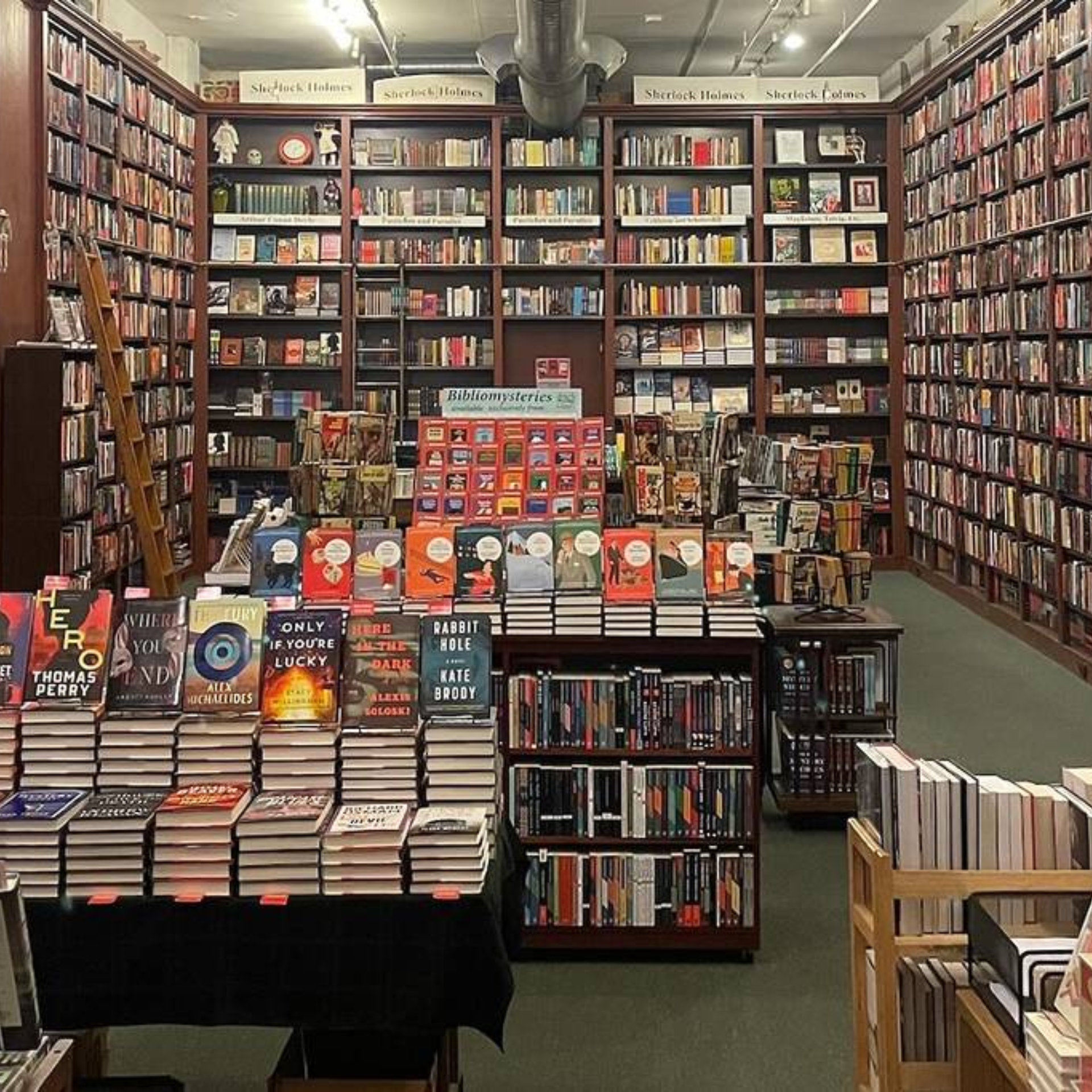 Bookstore interior with tall shelves labeled Sherlock Holmes, central tables stacked with mystery novels including Eve by Thomas Perry and Rabbit Hole by Kate Brody, Bibliomysteries display, wooden ladder, and cozy organized atmosphere