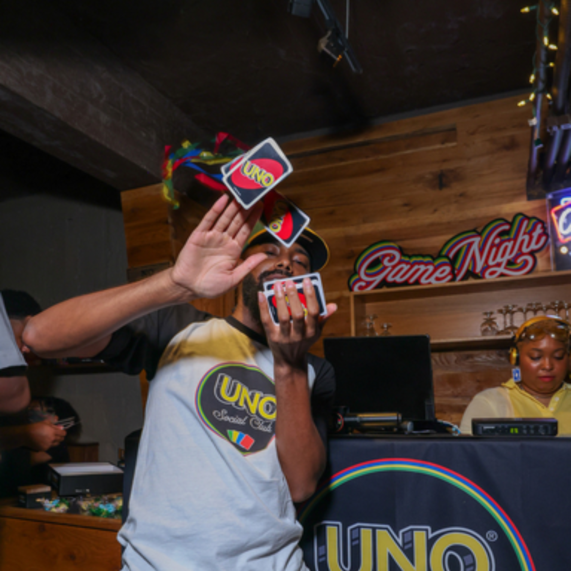 A man in an UNO Social Club shirt playfully shows off a hand of UNO playing cards at a neon-lit bar game night event