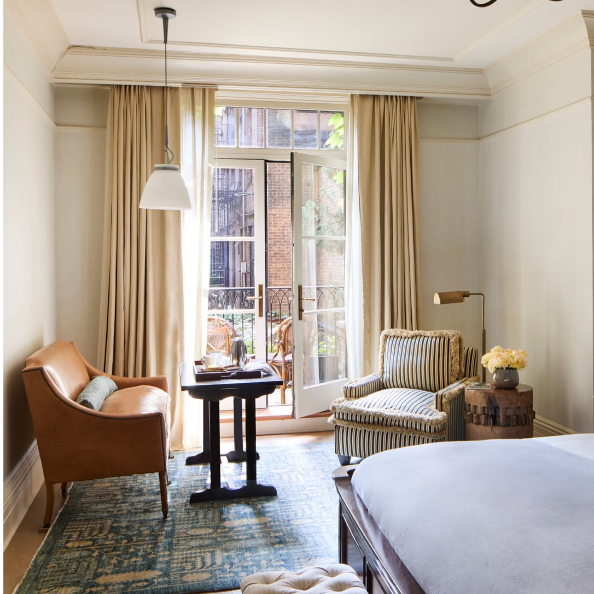 Elegant bedroom with tan leather armchair, striped chair, dark wood table, pendant light, French doors to balcony with wicker seating, patterned blue rug, tufted bench, brass lamp, and vase of yellow flowers