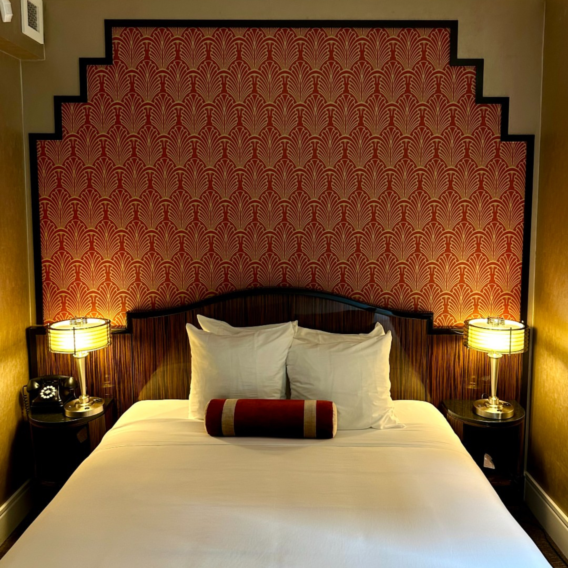 Cozy art deco bedroom with white sheets, red striped bolster pillow, dark wood stepped headboard, red patterned wallpaper, vintage rotary phone, and warm bedside lamps