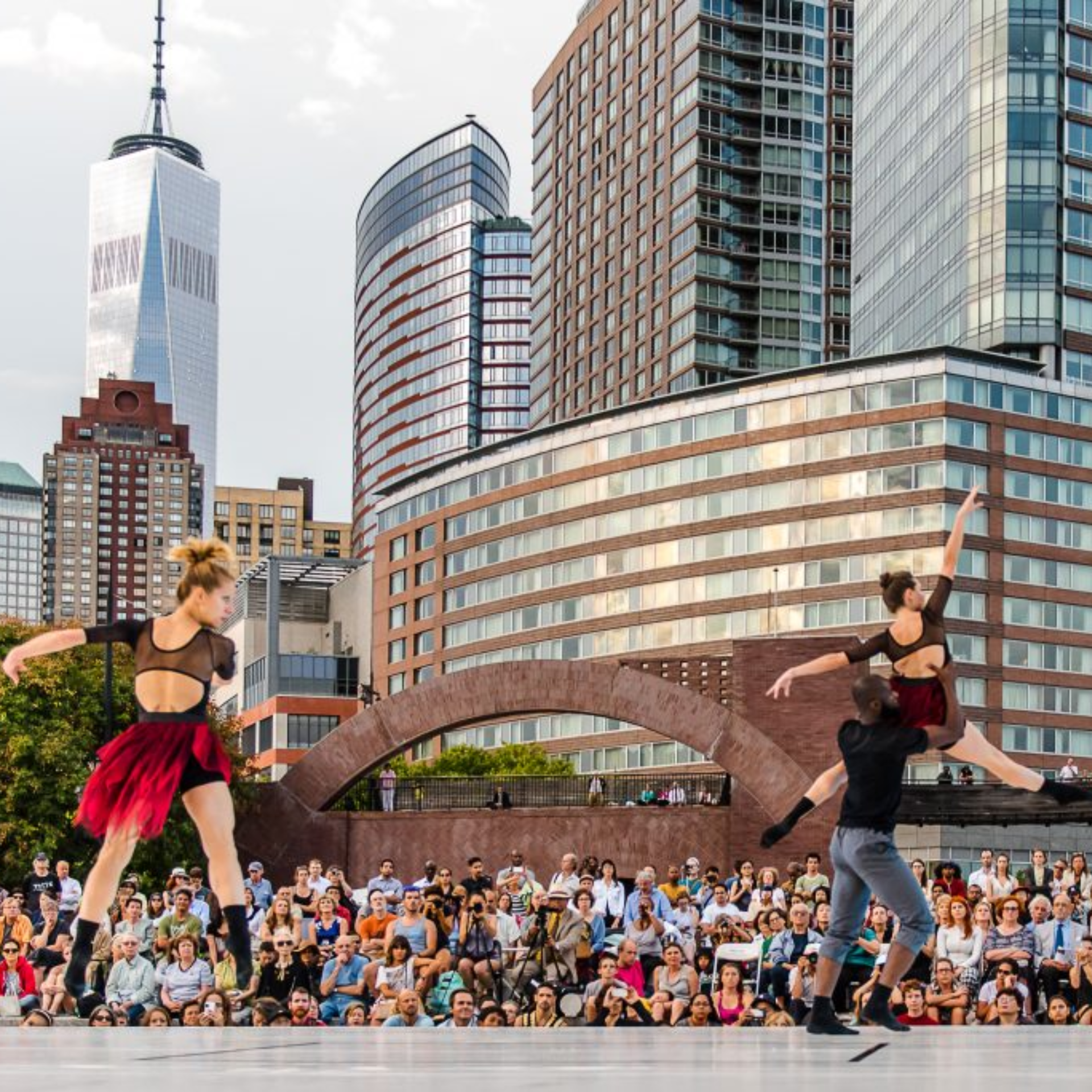 Outdoor NYC dance performance with three dancers in red skirts and black tops on stage before large audience, One World Trade Center skyline backdrop, cultural arts event featured in daily things to do in NYC any day