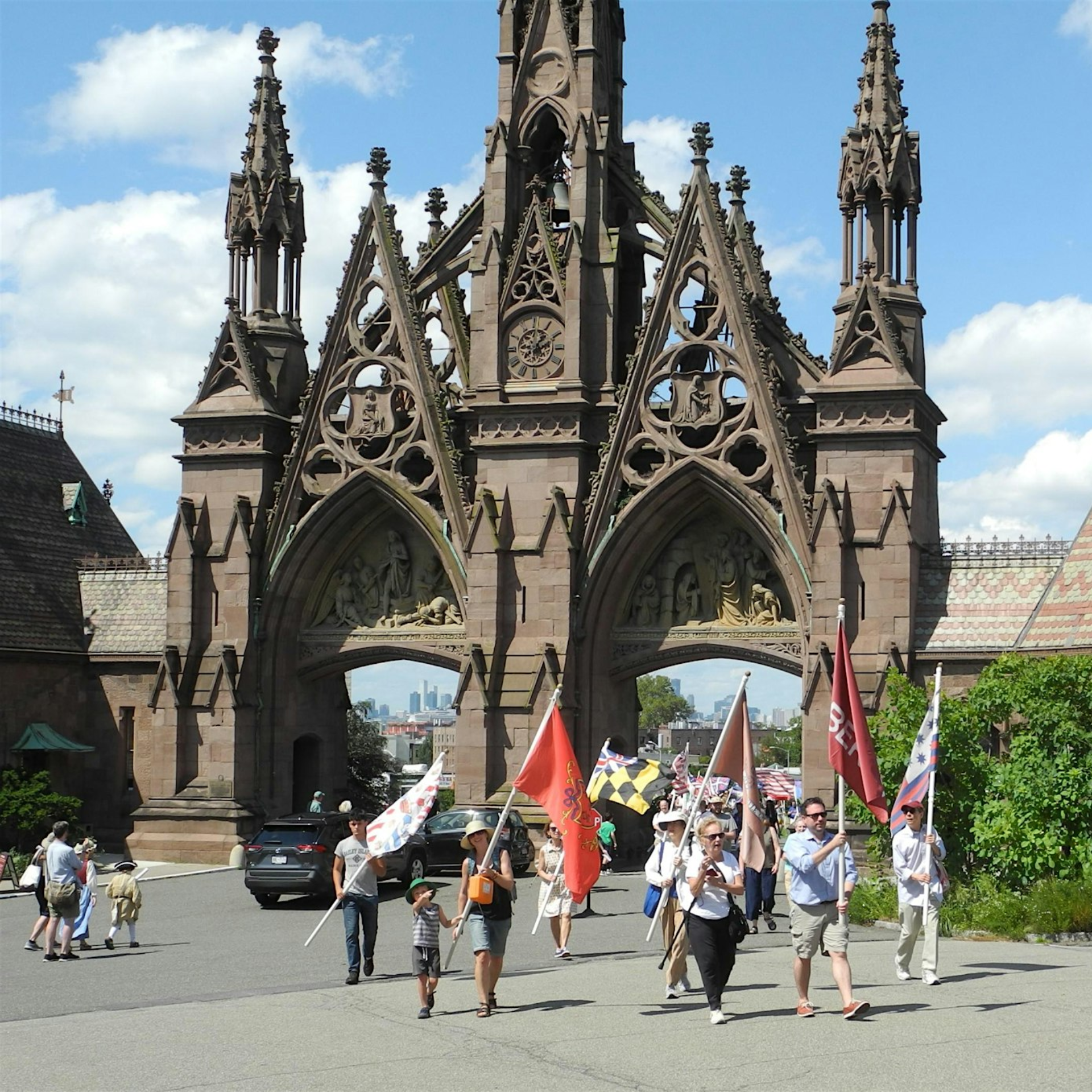 Parade with colorful flags in front of ornate Gothic stone archway and city skyline, NYC cultural procession featured in daily things to do in NYC any day