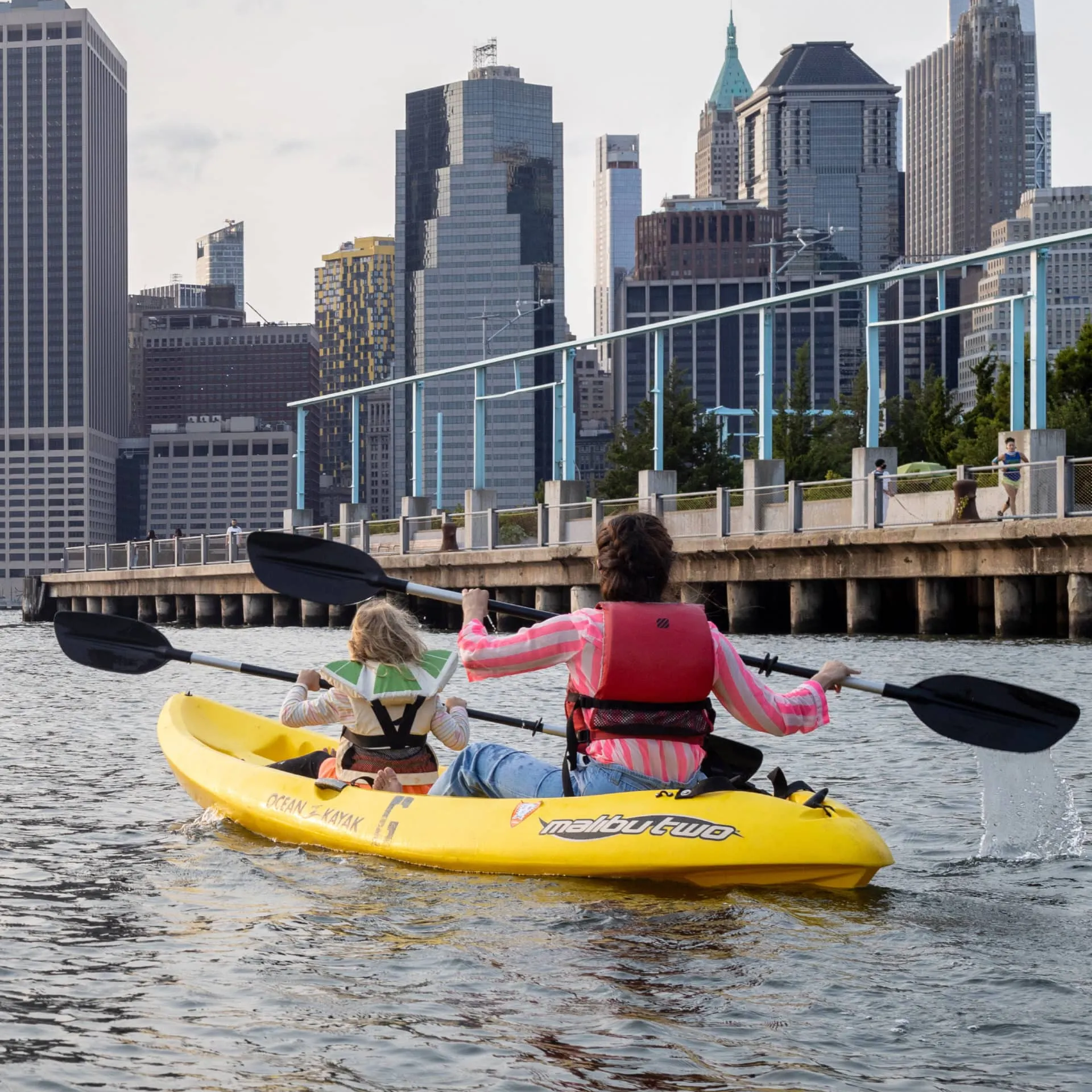 Two people in yellow tandem kayak labeled Malibu Two paddling near urban waterfront pier with city skyline of tall buildings in background