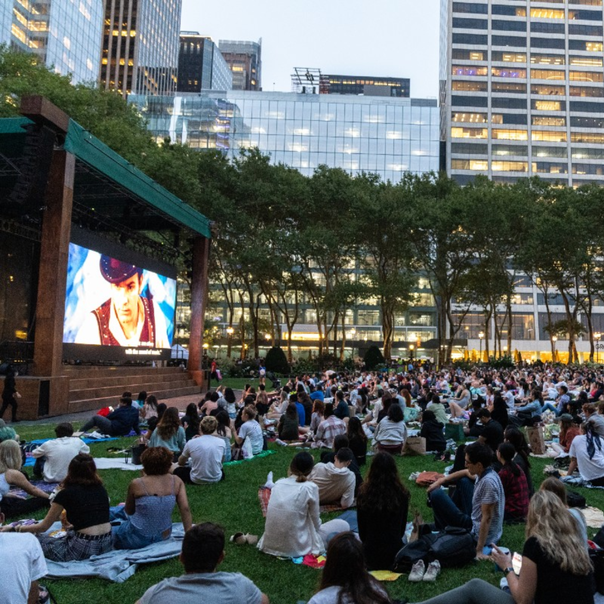 NYC outdoor movie screening in city park with crowd on blankets watching film under tall glass buildings, communal urban cinema featured in daily things to do in NYC any day