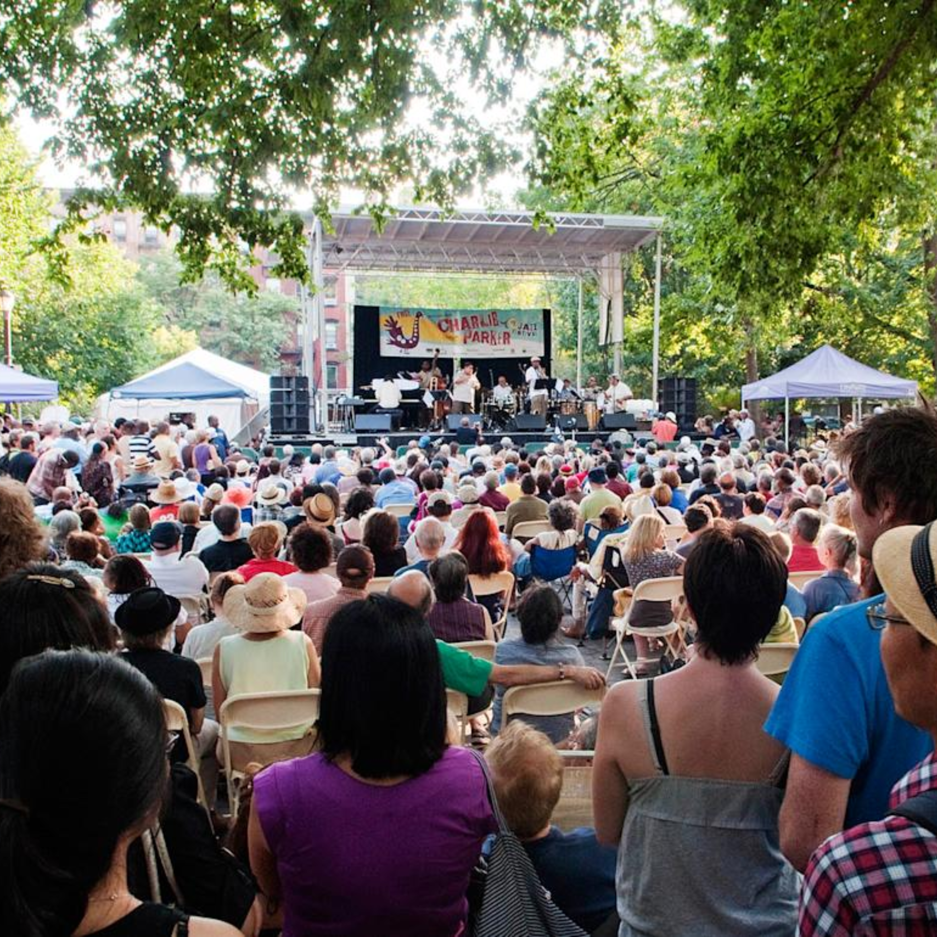 NYC park jazz festival with Charlie Parker tribute stage, band performing for large seated crowd under trees, cultural music gathering featured in daily things to do in NYC any day
