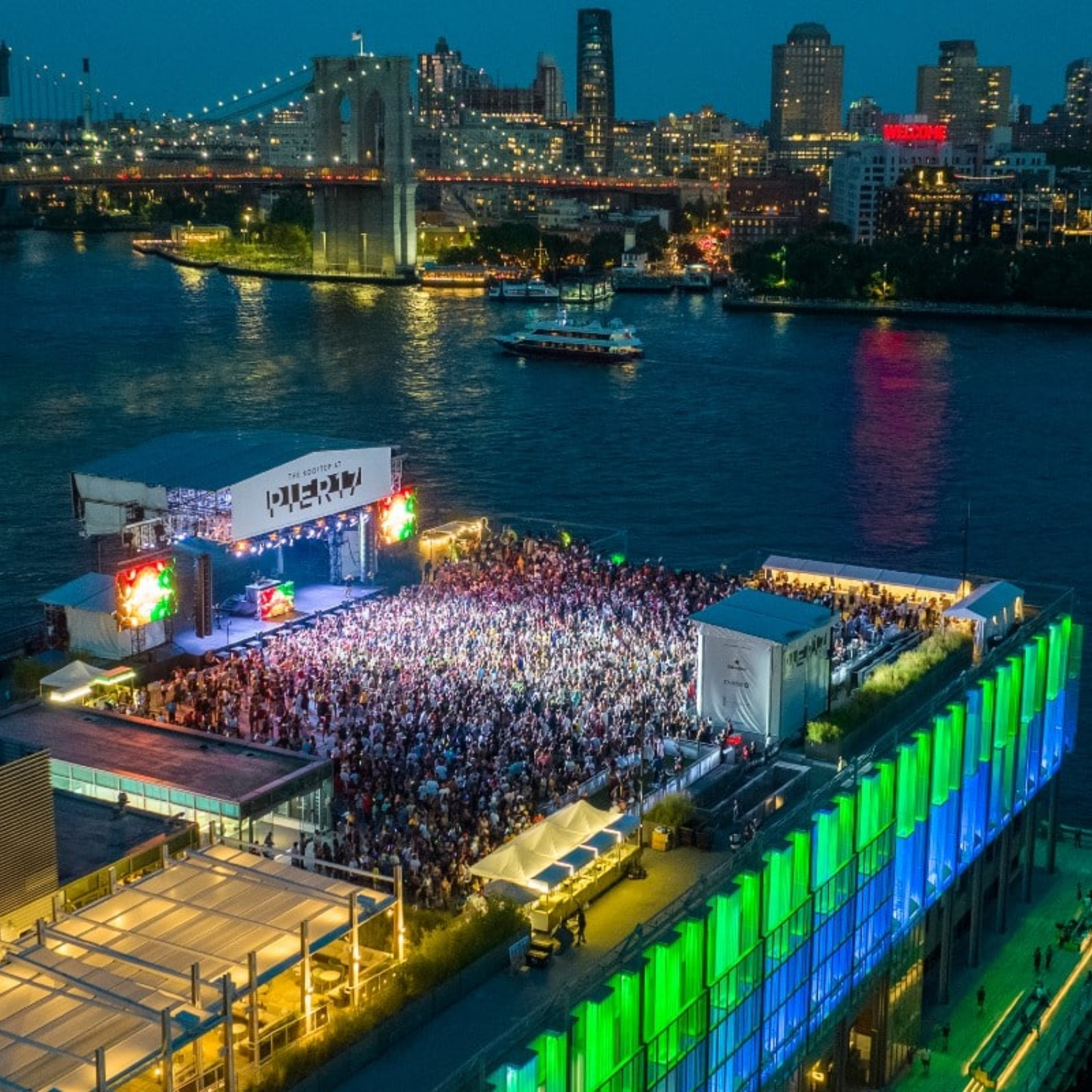 Pier 17 NYC rooftop concert with dense crowd, colorful stage lights, Brooklyn Bridge and Manhattan skyline at night, vibrant urban nightlife featured in daily things to do in NYC any day